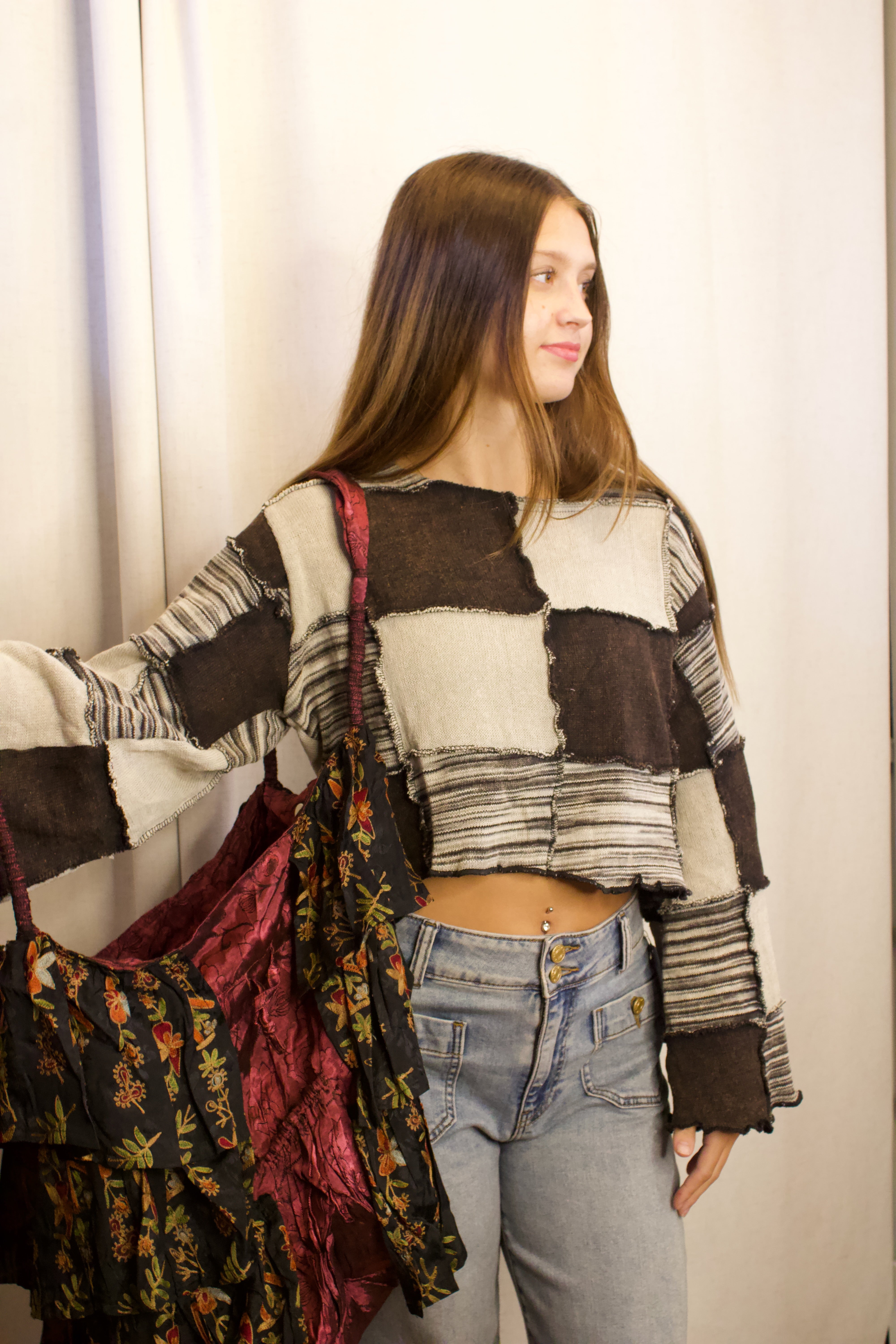 A young woman wearing a brown, beige, and black patchwork long-sleeve crop top and light wash jeans, holding a large dark floral patterned tote bag against a neutral off-white curtain background.