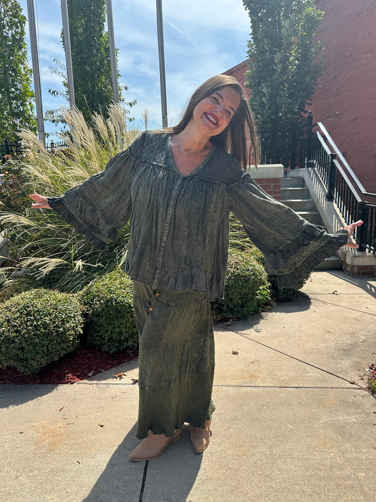 A woman wearing a matching olive green long-sleeve button-up shirt and wide-leg skirt stands outdoors on a concrete walkway with her arms outstretched, smiling broadly.