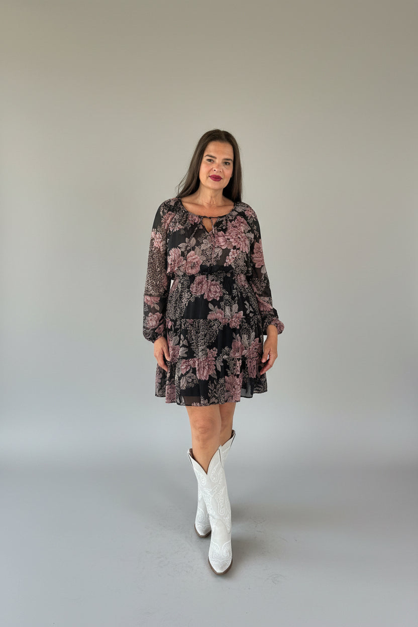 A woman stands wearing a black long-sleeve mini dress with a pink floral pattern and white cowboy boots.