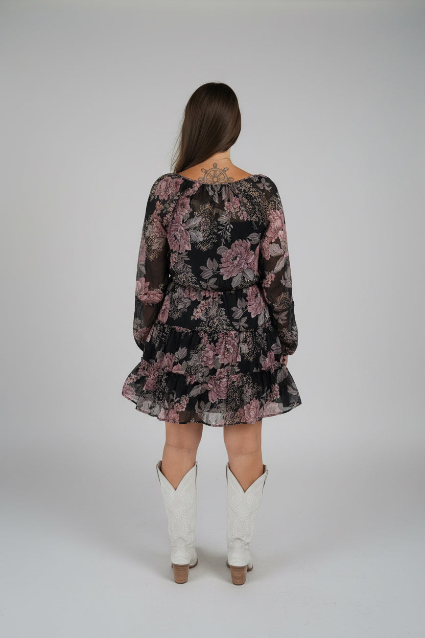 A woman viewed from behind wearing a black long-sleeve mini dress with a dusty rose floral pattern, paired with white cowboy boots.