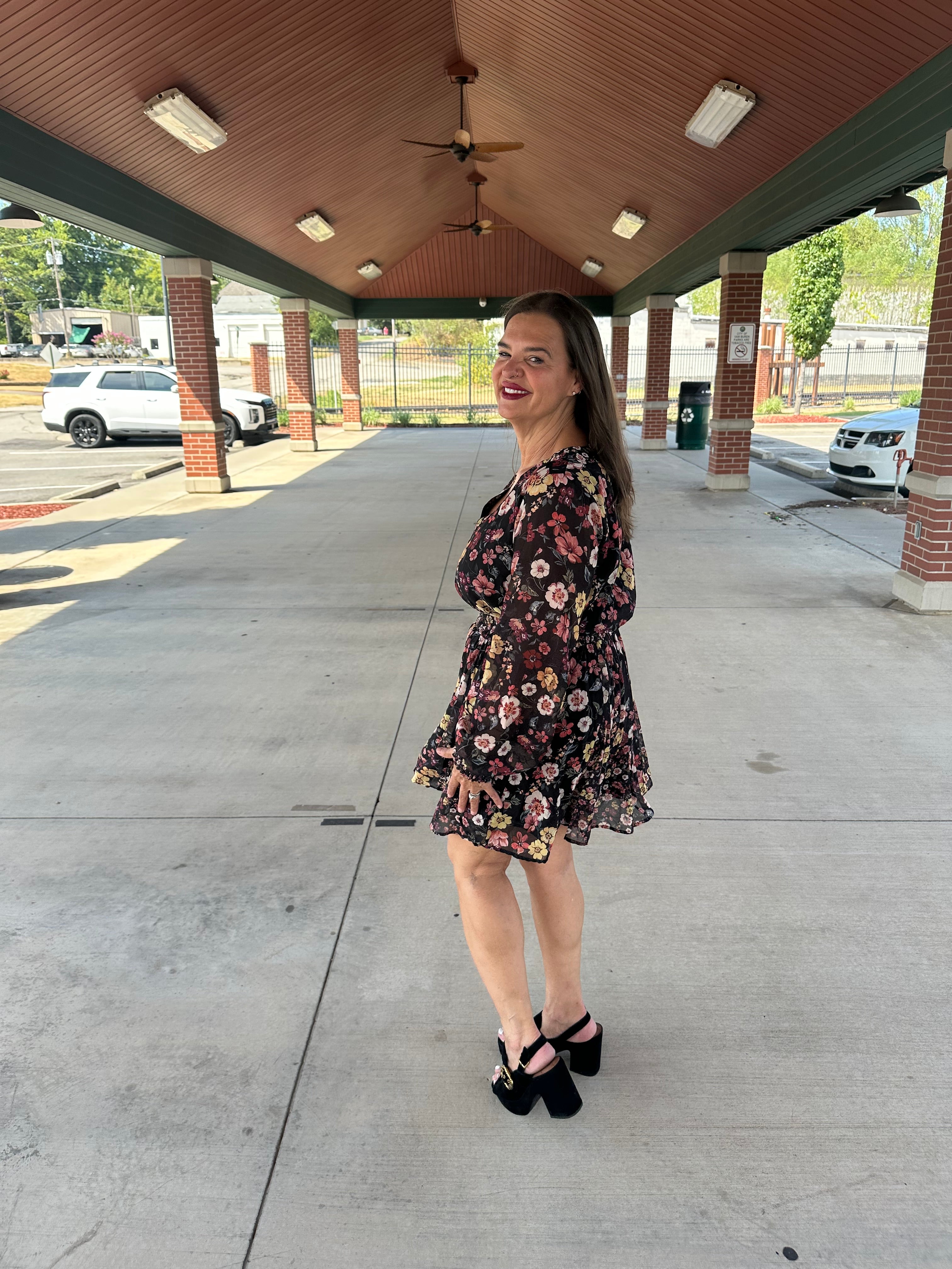 A woman wearing a short black floral dress and black high heels stands under a covered walkway, looking back over her shoulder.
