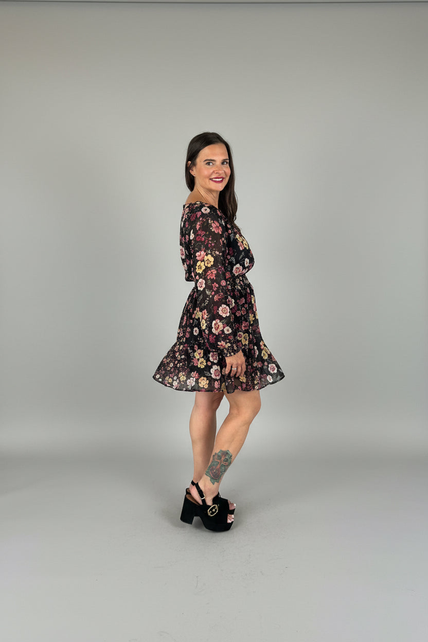 Woman smiling over shoulder in Smocked Waist V-Neck Mini Dress in black floral print, side profile showing smocked cuffs and pleated hem, black platform heels