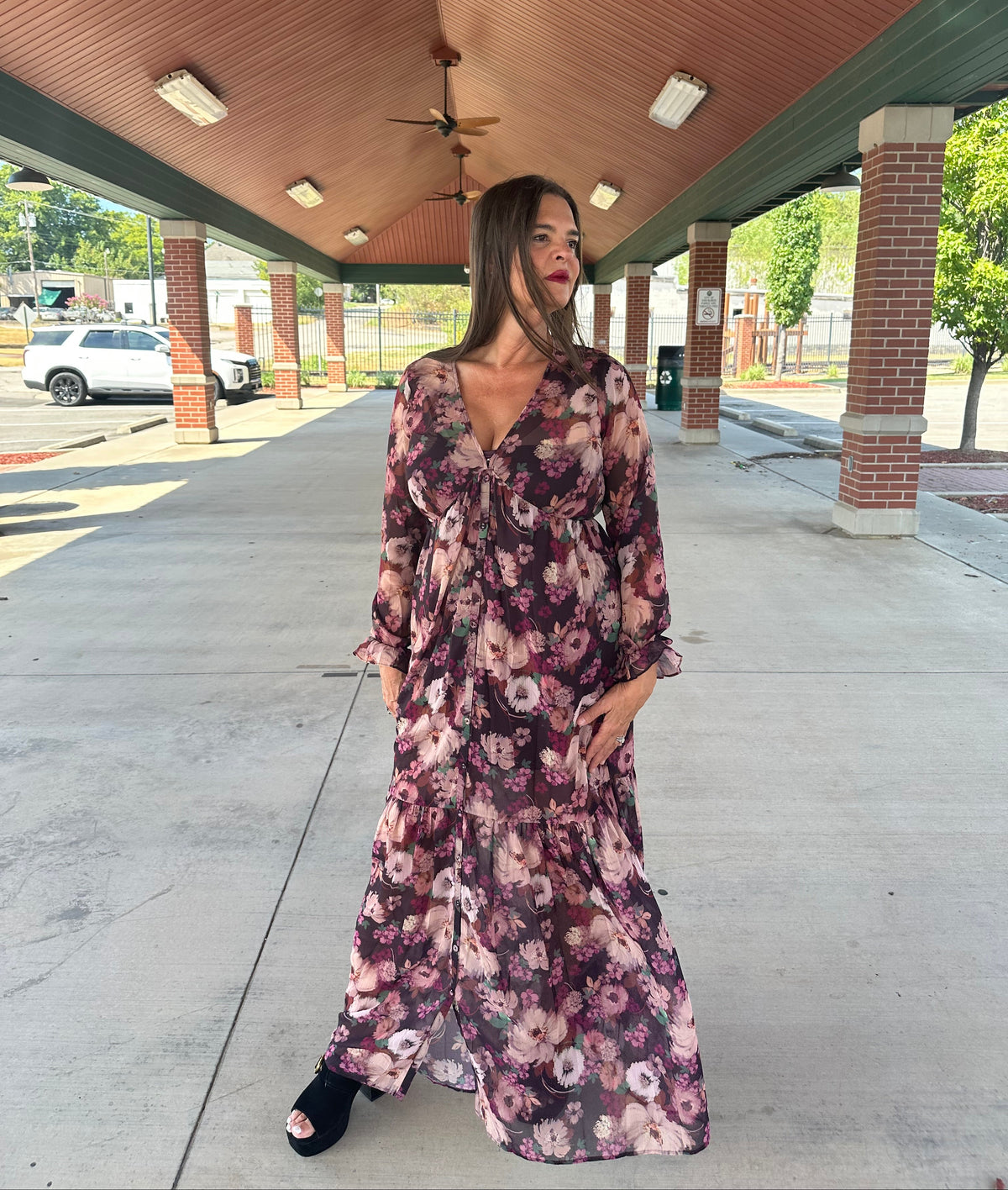 Woman wearing burgundy floral long-sleeve maxi dress with V-neckline and tiered hem, styled with black heeled sandals, outdoor pavilion setting