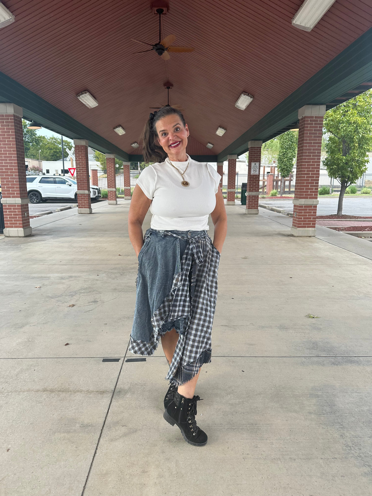 A woman posing under a covered walkway wearing a white t-shirt, a denim skirt, and a black-and-white gingham shirt tied around her waist, paired with black lace-up boots.