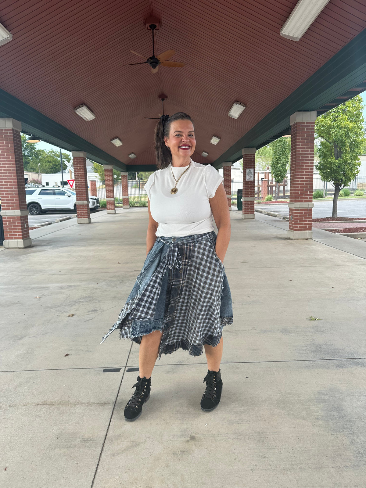 A woman stands under a covered walkway wearing a white short-sleeve top, a blue and white gingham skirt tied at the waist, and black lace-up boots.