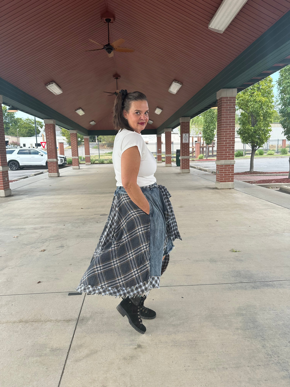 A woman posing under a covered walkway wearing a white short-sleeve t-shirt, a long black and white plaid skirt, a blue denim shirt tied around her waist, and black lace-up boots.