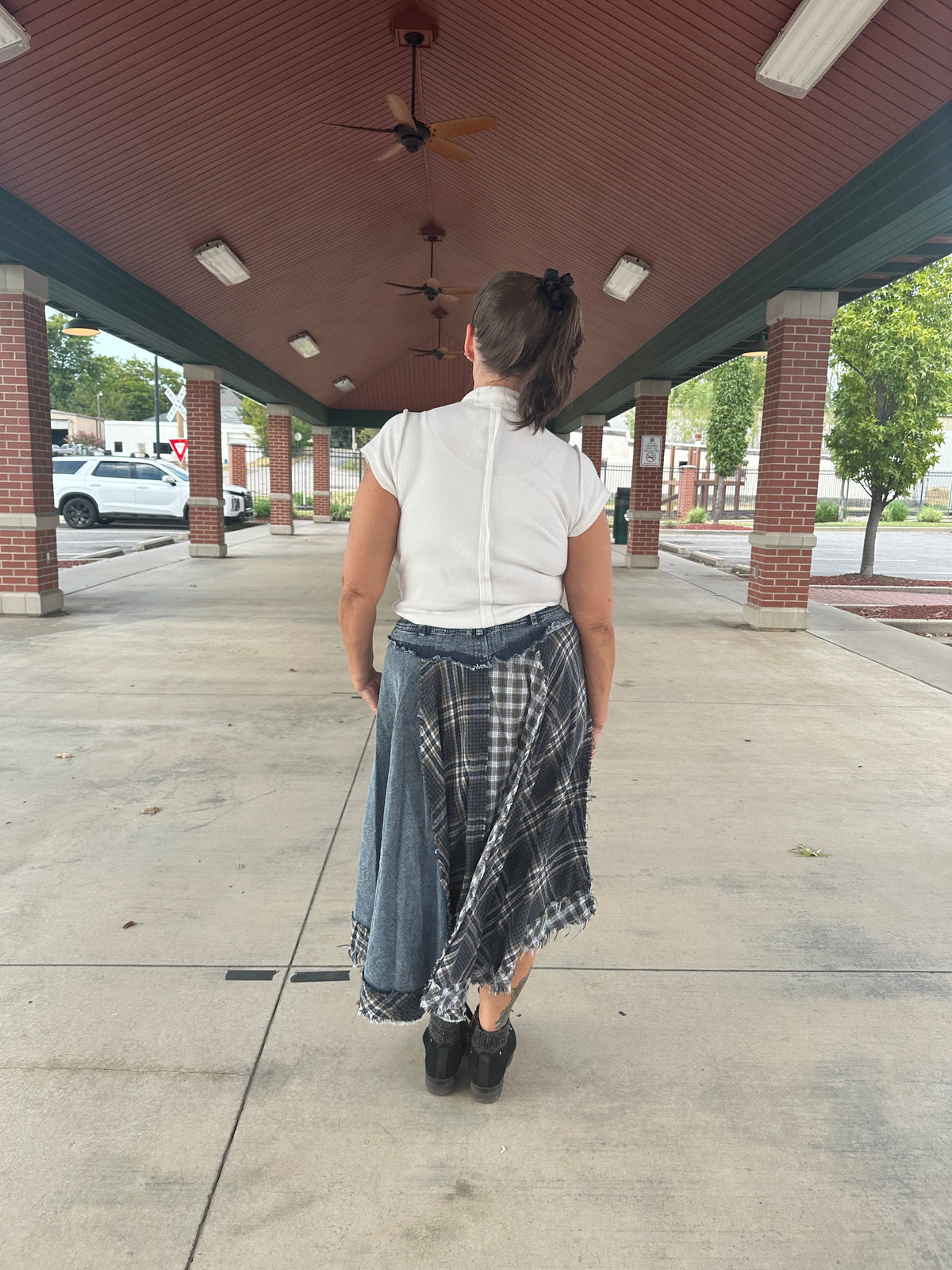 A woman viewed from behind wearing a white short-sleeve top and a long denim skirt with a black-and-white plaid shirt tied around her waist, standing on a covered outdoor walkway.