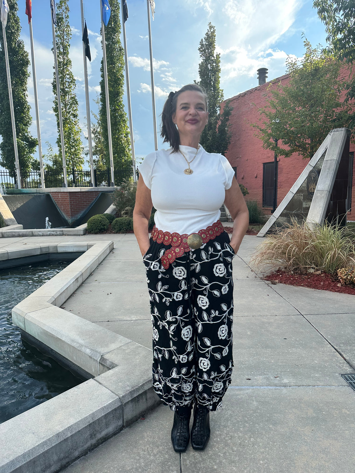 A woman stands outdoors on a concrete plaza next to a water feature, wearing a white short-sleeve top, black wide-leg pants with a white floral pattern and a red patterned belt, and black ankle boots.