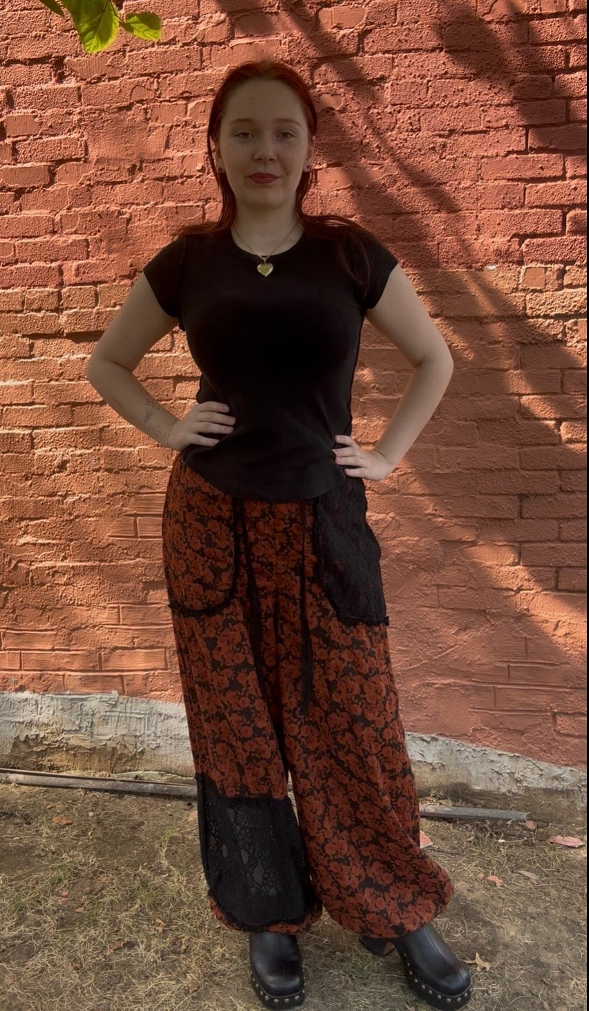 A woman with red hair stands in front of a red brick wall, wearing a black short-sleeve top, wide-leg patterned pants in red and black, and black boots.
