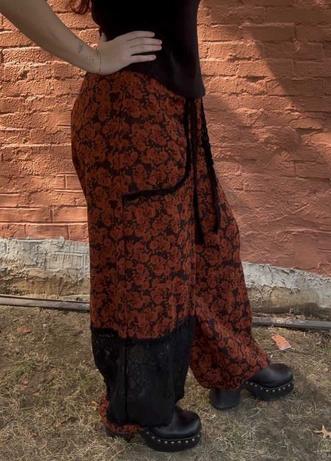 A person wearing loose, wide-leg patterned pants in rust orange and black, paired with a black top and black platform boots, standing outdoors against a terracotta brick wall.