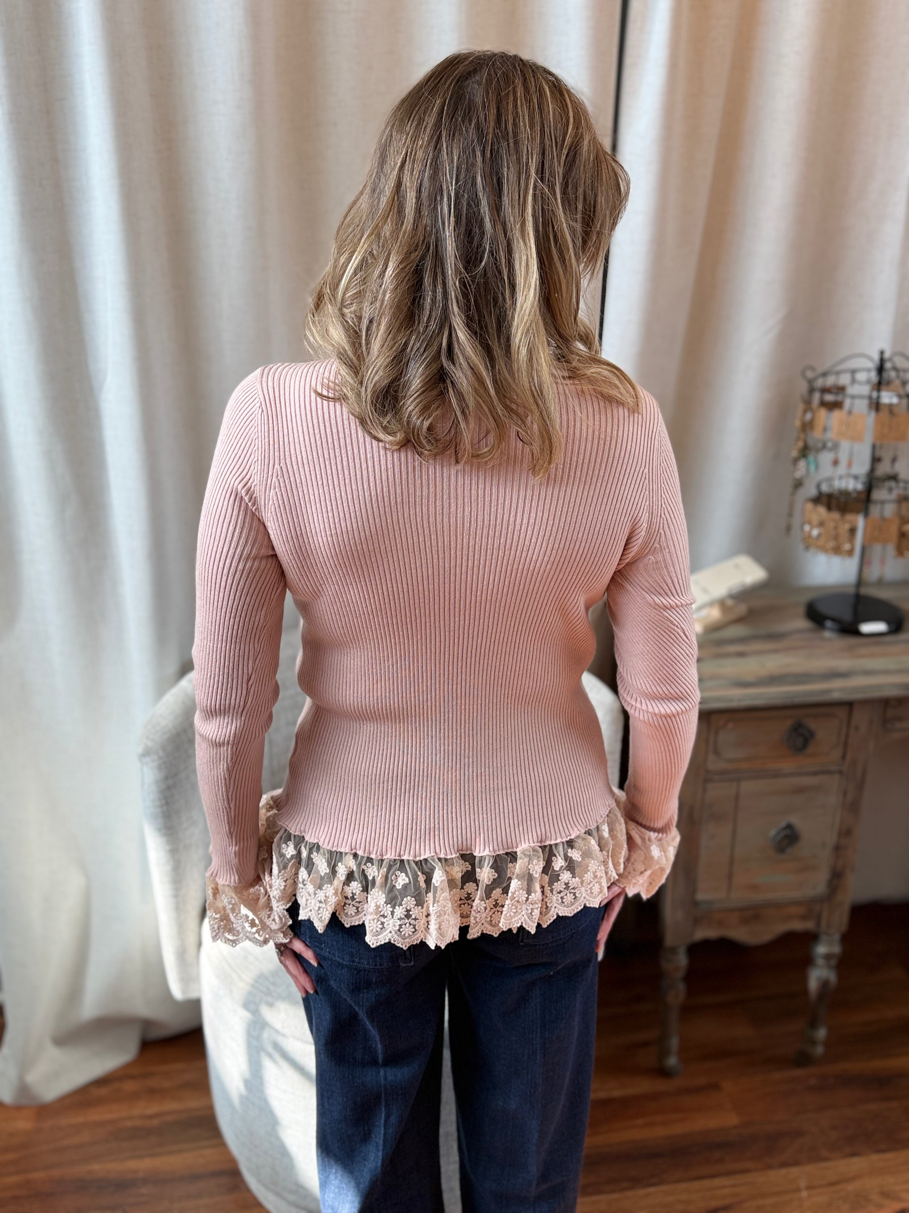 Person wearing a pink sweater with lace trim in a room with curtains and a desk.
