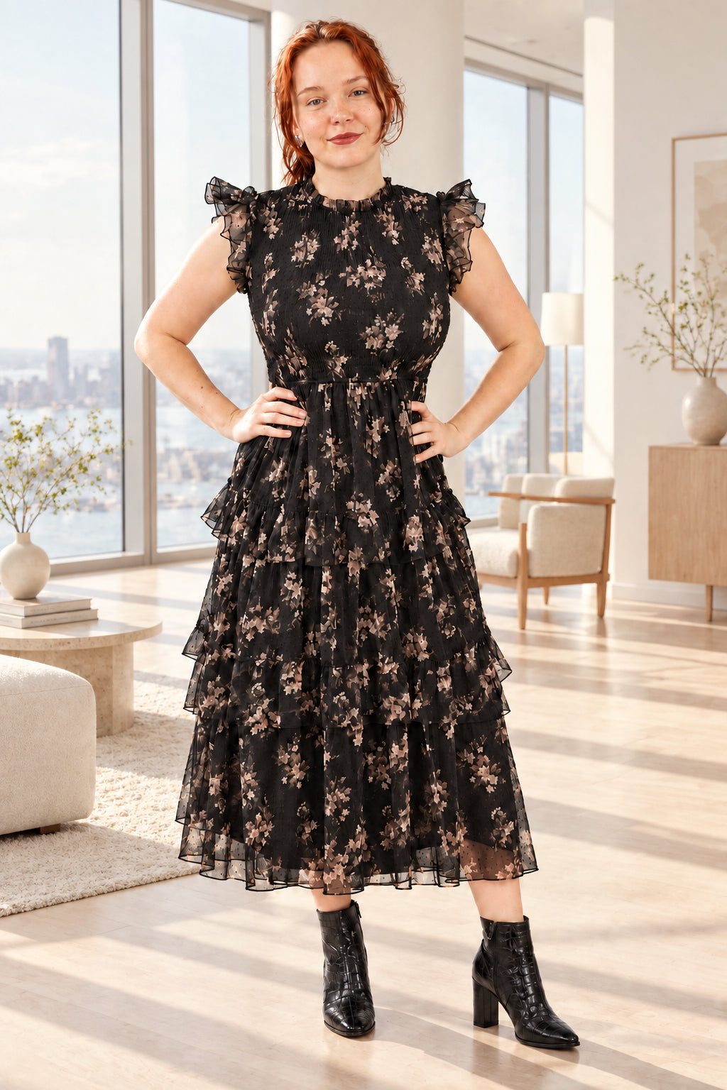 A woman with red hair wearing a black floral tulle midi dress with ruffled cap sleeves and black ankle boots, standing in a living room.
