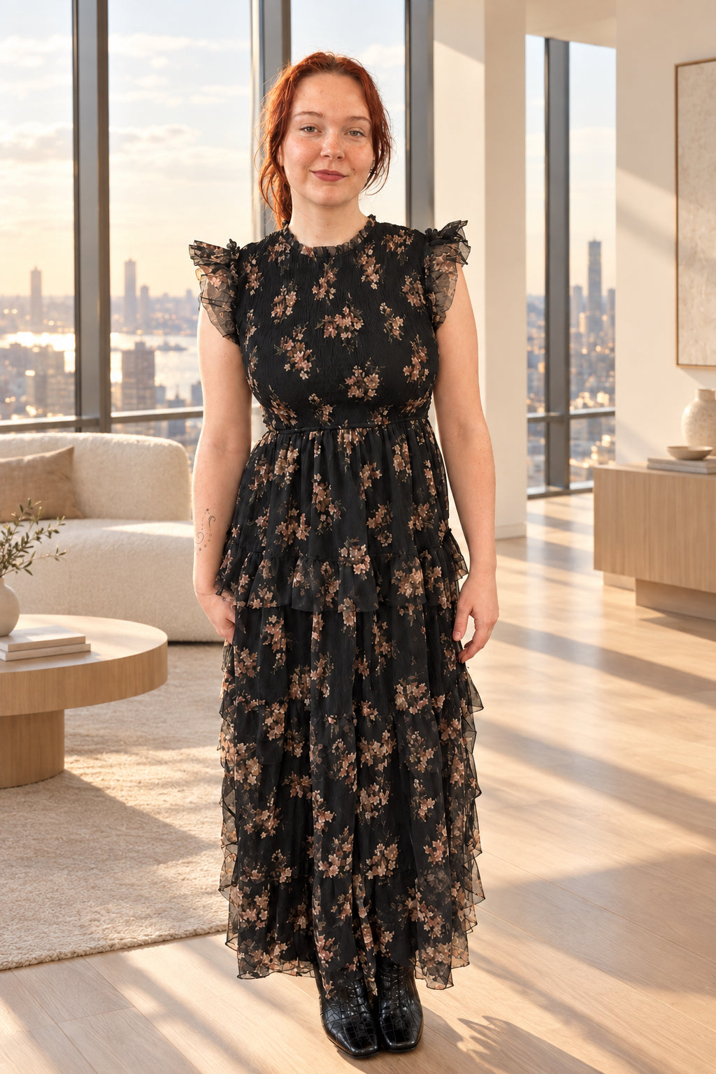 A woman with red hair wearing a black floral midi dress with sheer flutter sleeves and a ruffled hem, standing in a living room.