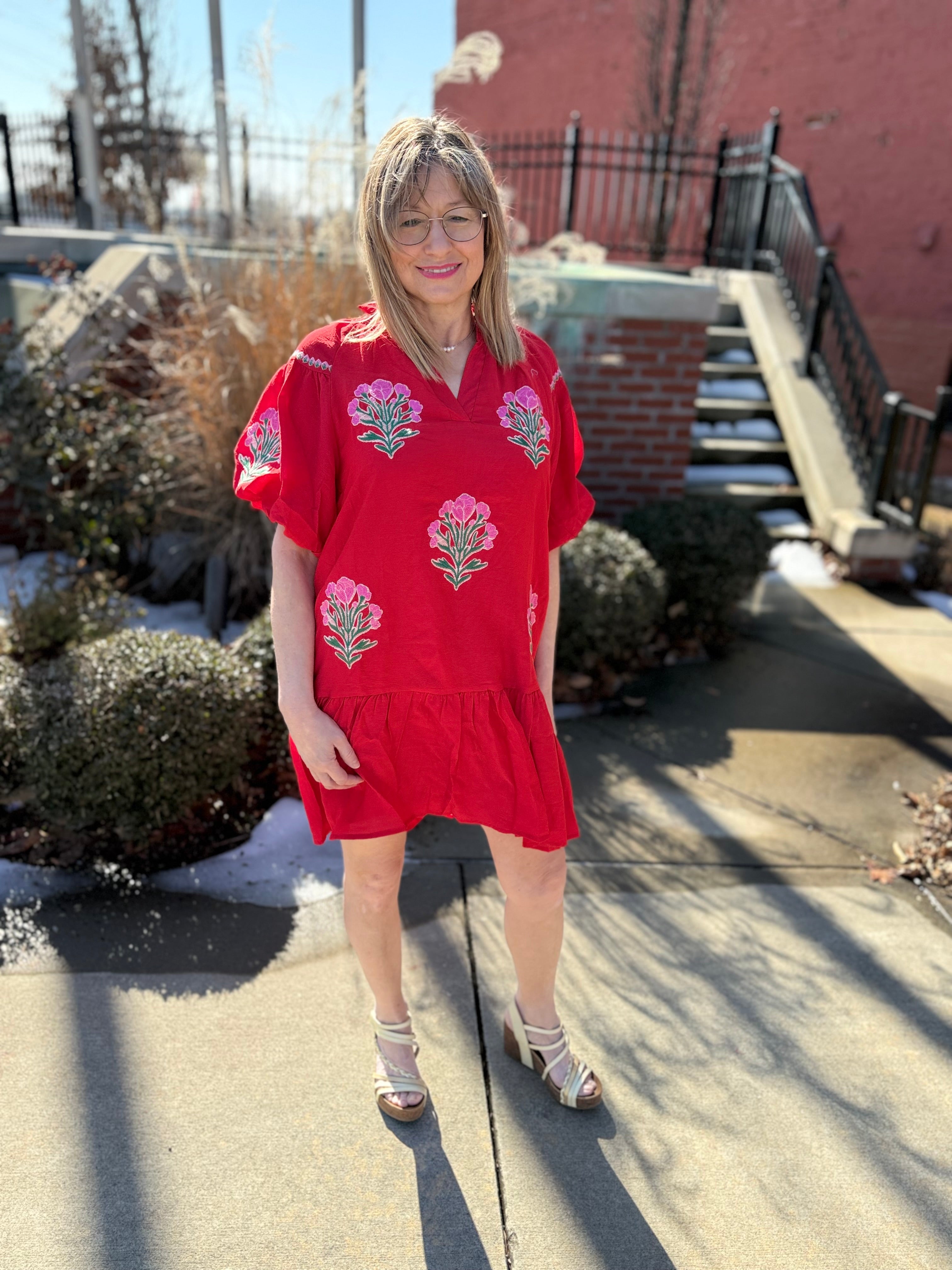 Woman wearing a red dress with floral patterns standing outdoors on a sunny day.