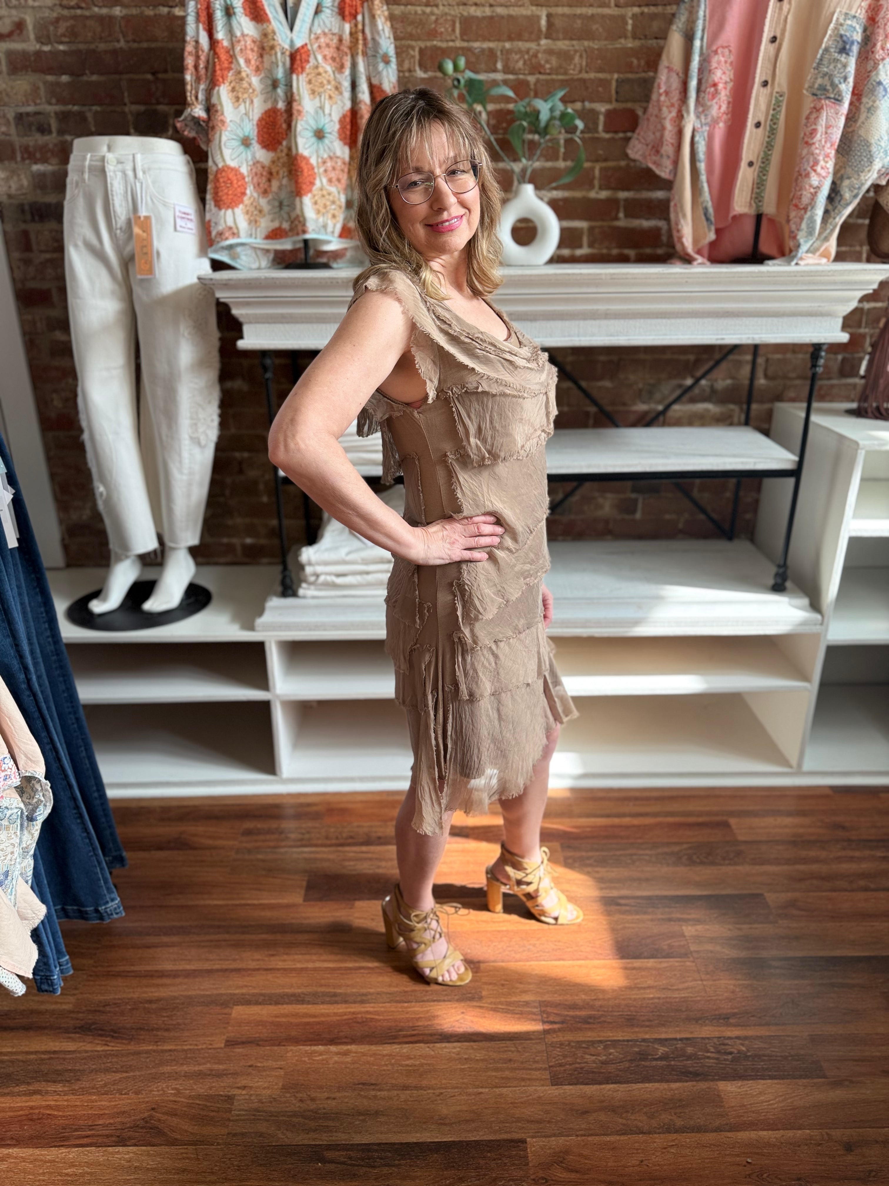 Woman in a beige dress standing in a clothing store with various outfits on display.