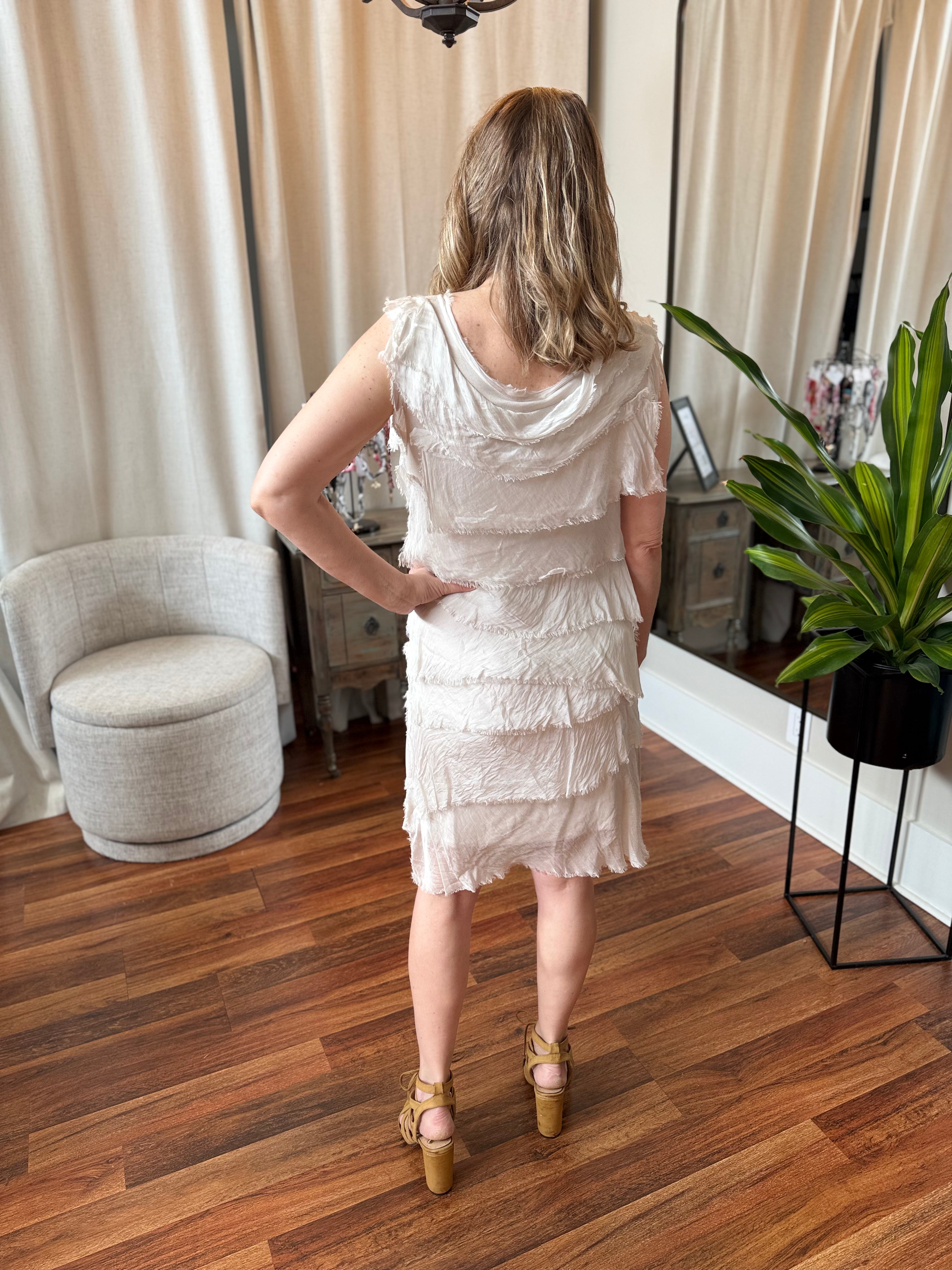 Woman in a white dress standing in a room with wooden flooring and curtains.