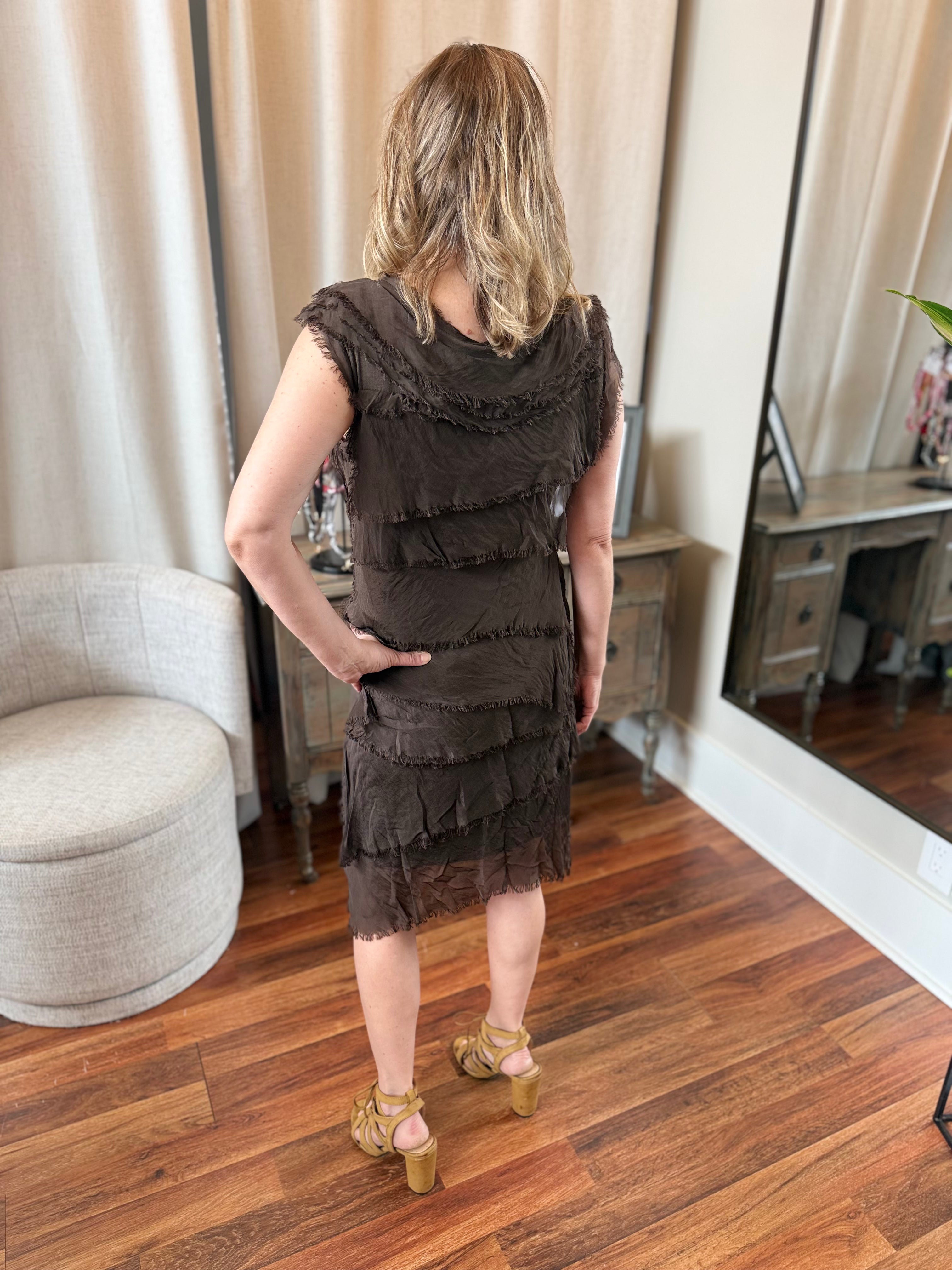 Woman wearing a brown dress standing in a room with wooden flooring and curtains.
