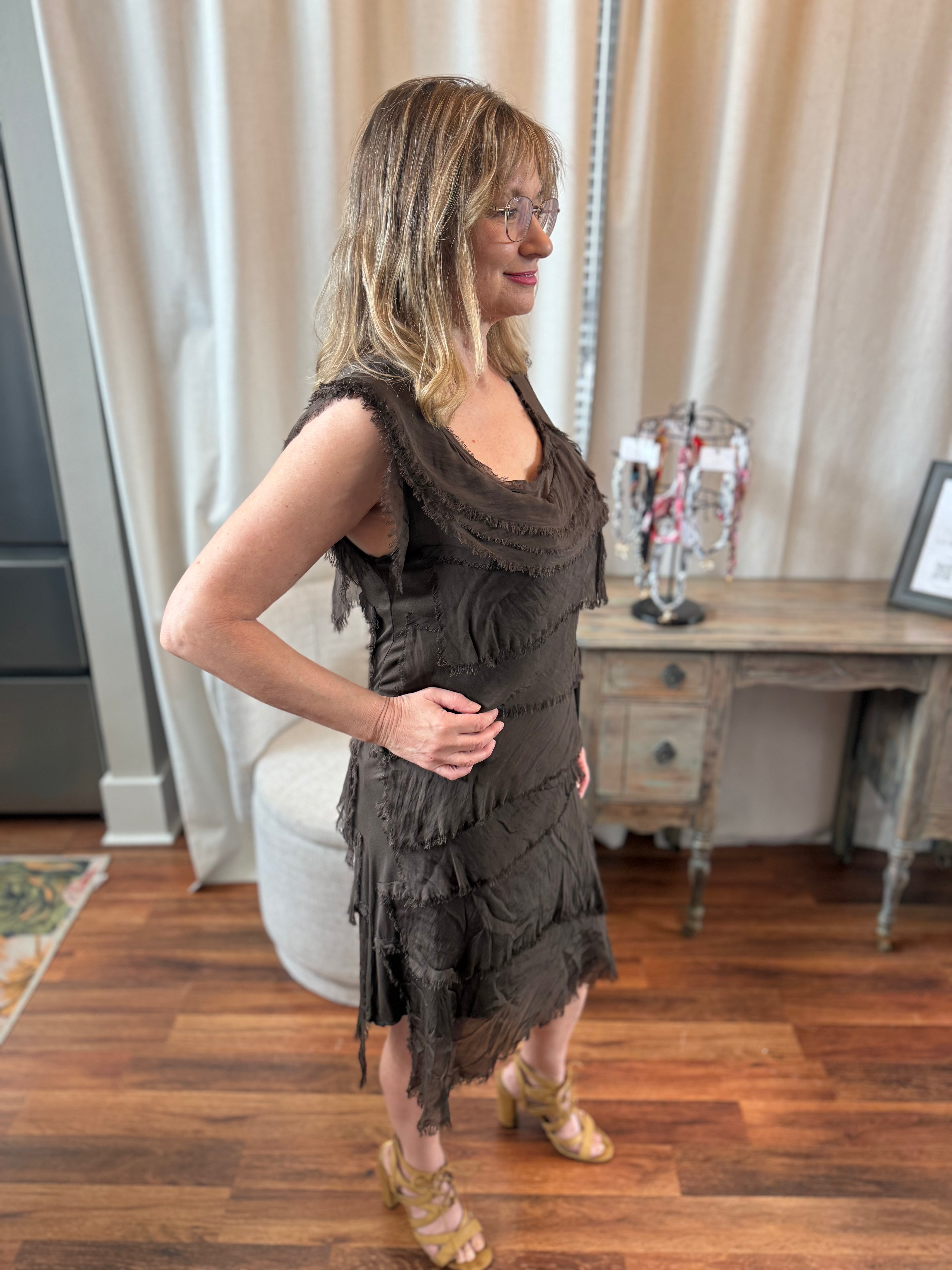 Woman wearing a brown fringed dress standing in a room with wooden flooring and a mirror.
