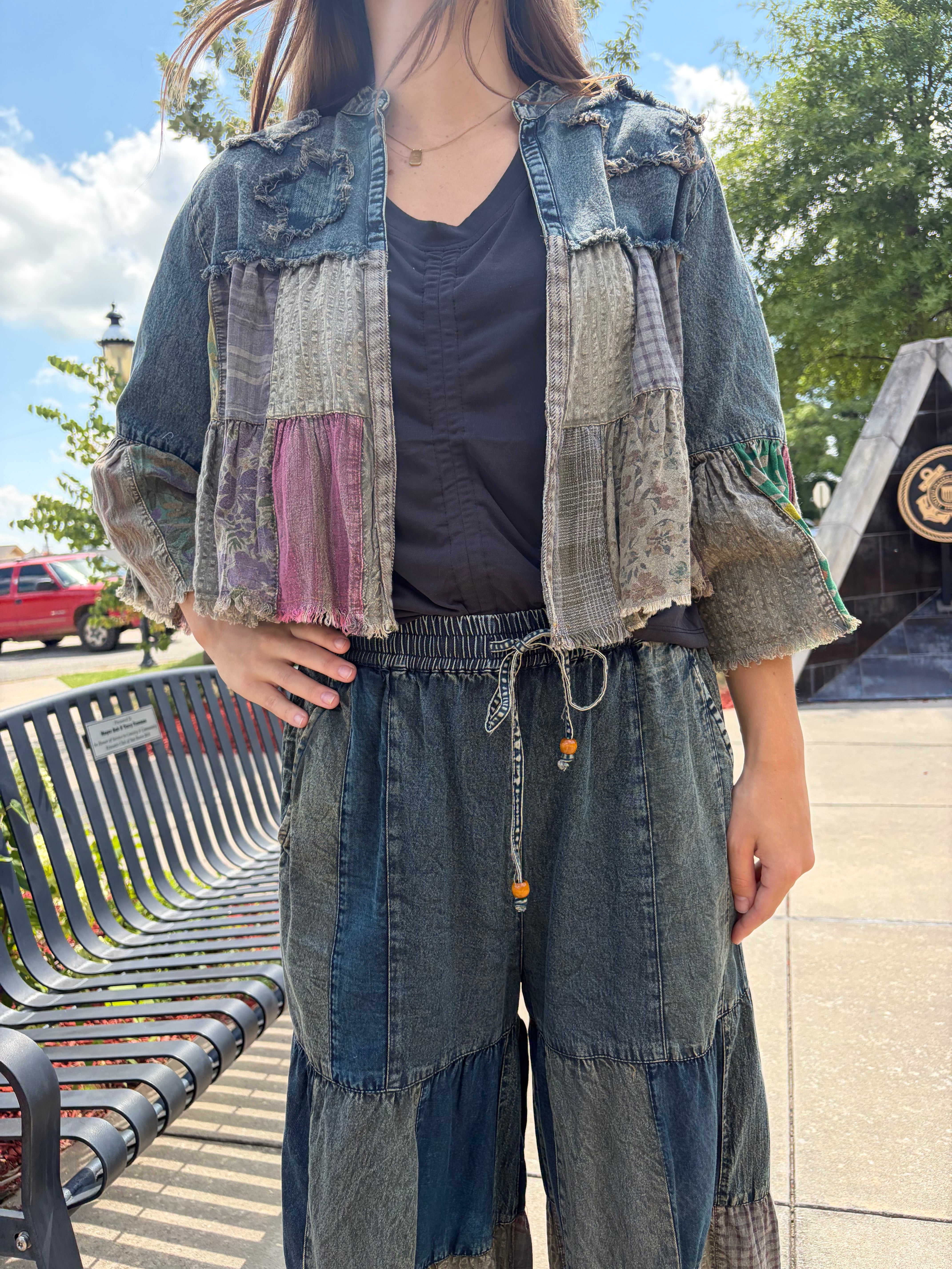 Front close-up of the cropped shrug open over a black tee, showing the mixed patchwork panels (lace, plaid, floral, pink) with frayed hem and ruffle sleeves