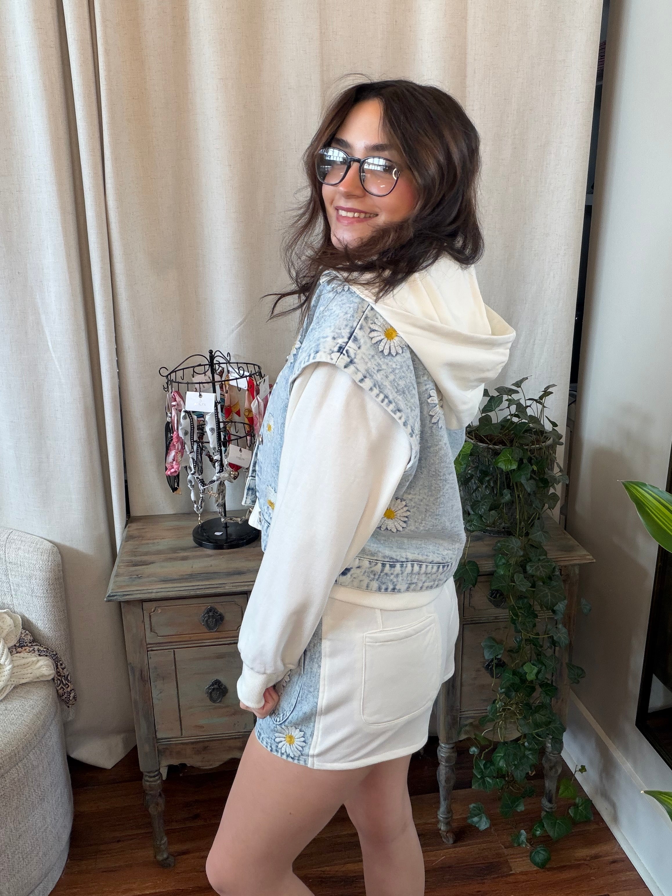 Woman wearing a denim vest over a white shirt and shorts in a room with curtains and a plant.