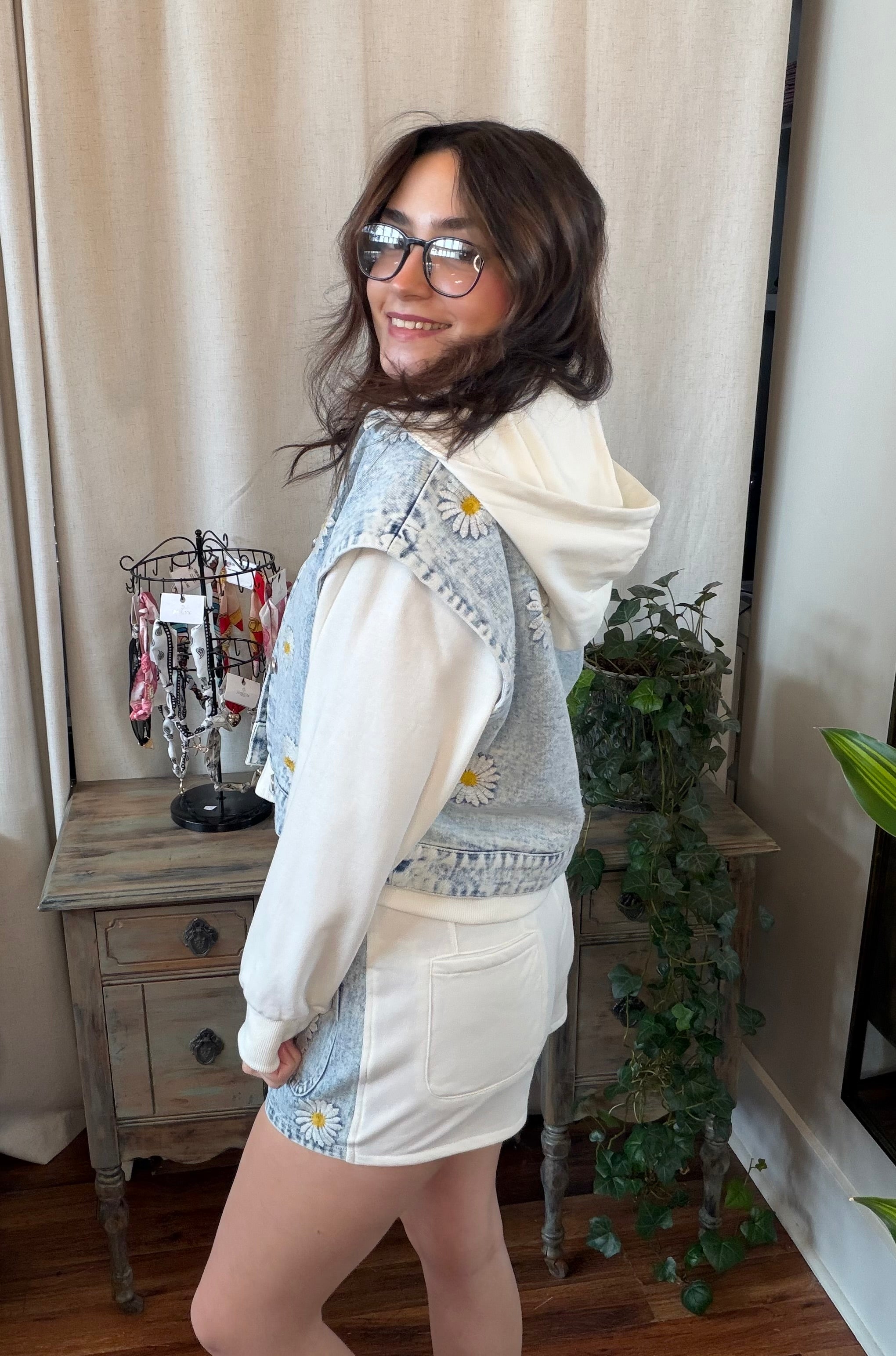 Woman wearing a white hoodie and denim jacket with floral patterns in a room.