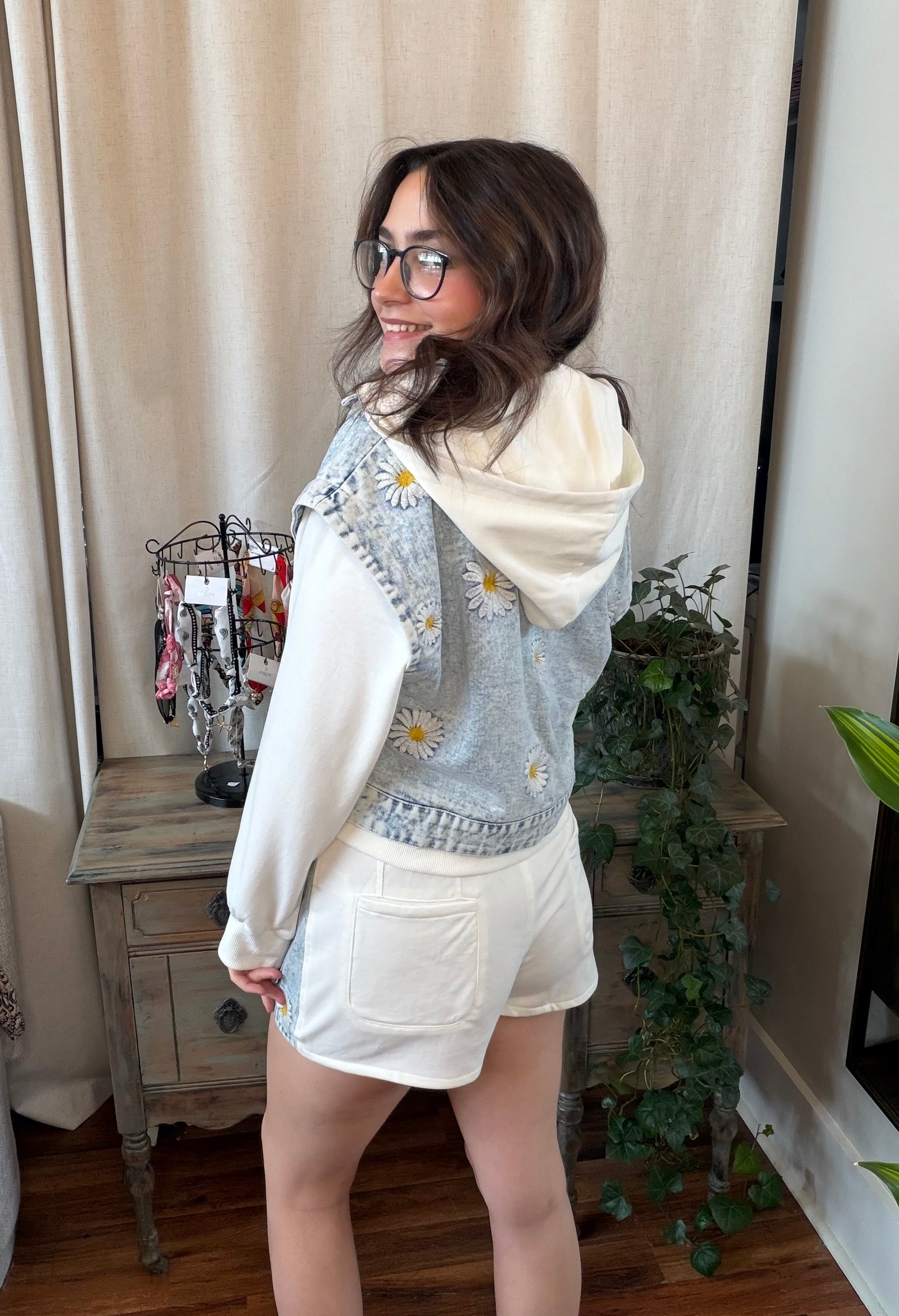 Person wearing a light blue floral cardigan over a white hoodie with white shorts, standing indoors.