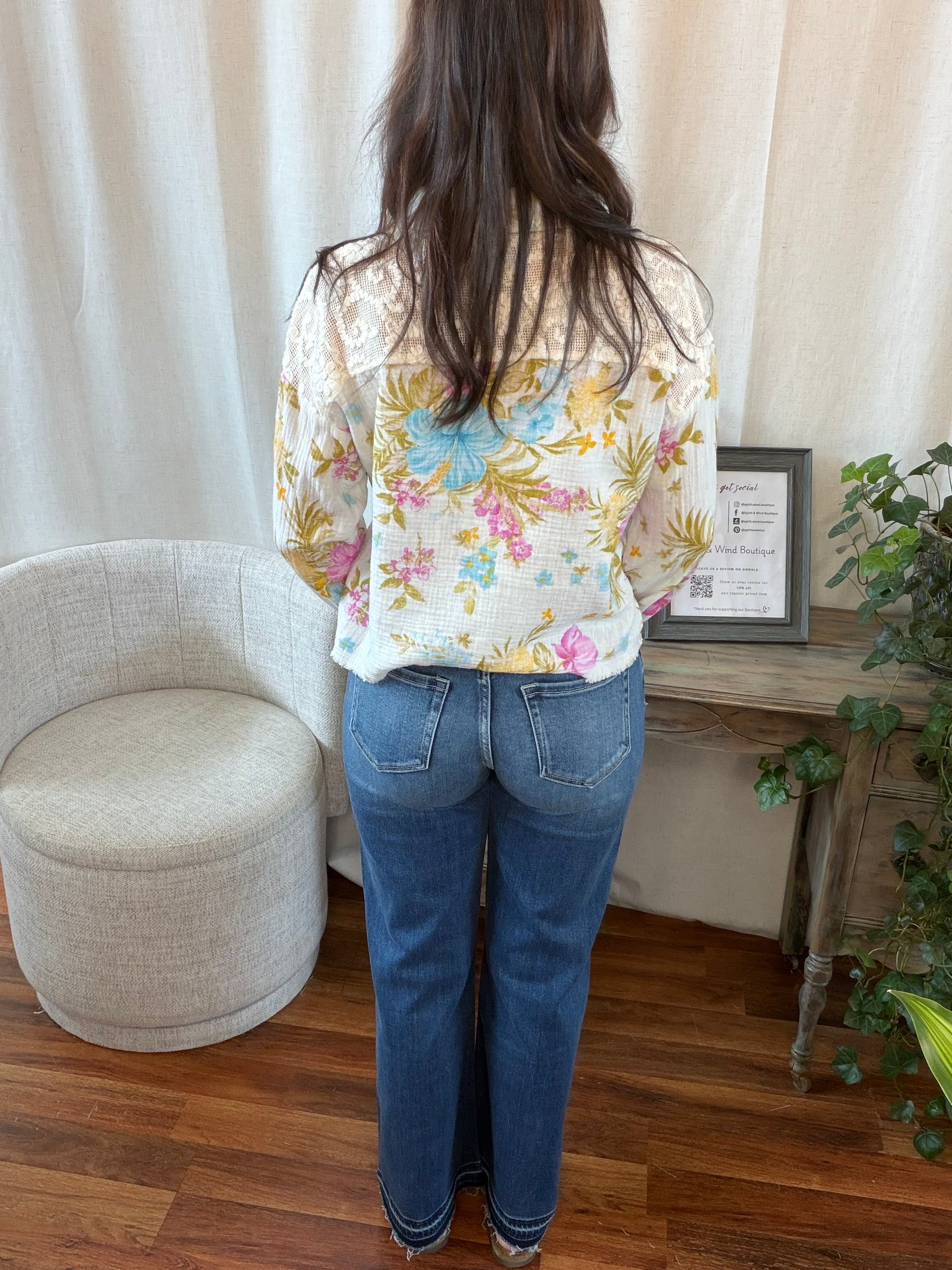 Person wearing a floral blouse and blue jeans standing in a room with a white curtain and wooden floor.
