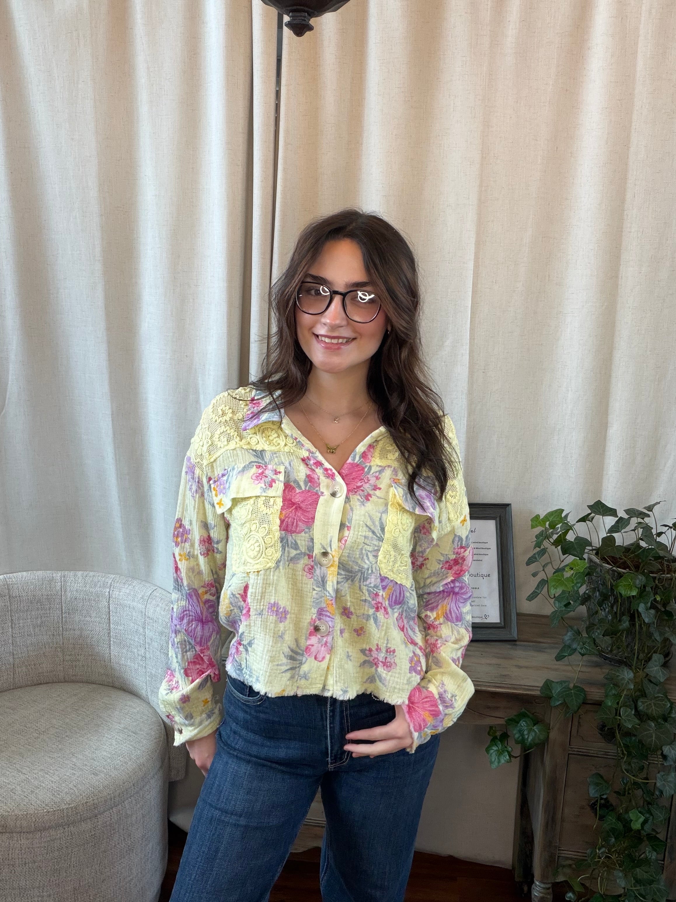 Woman wearing a floral blouse and glasses indoors with curtains and a plant in the background