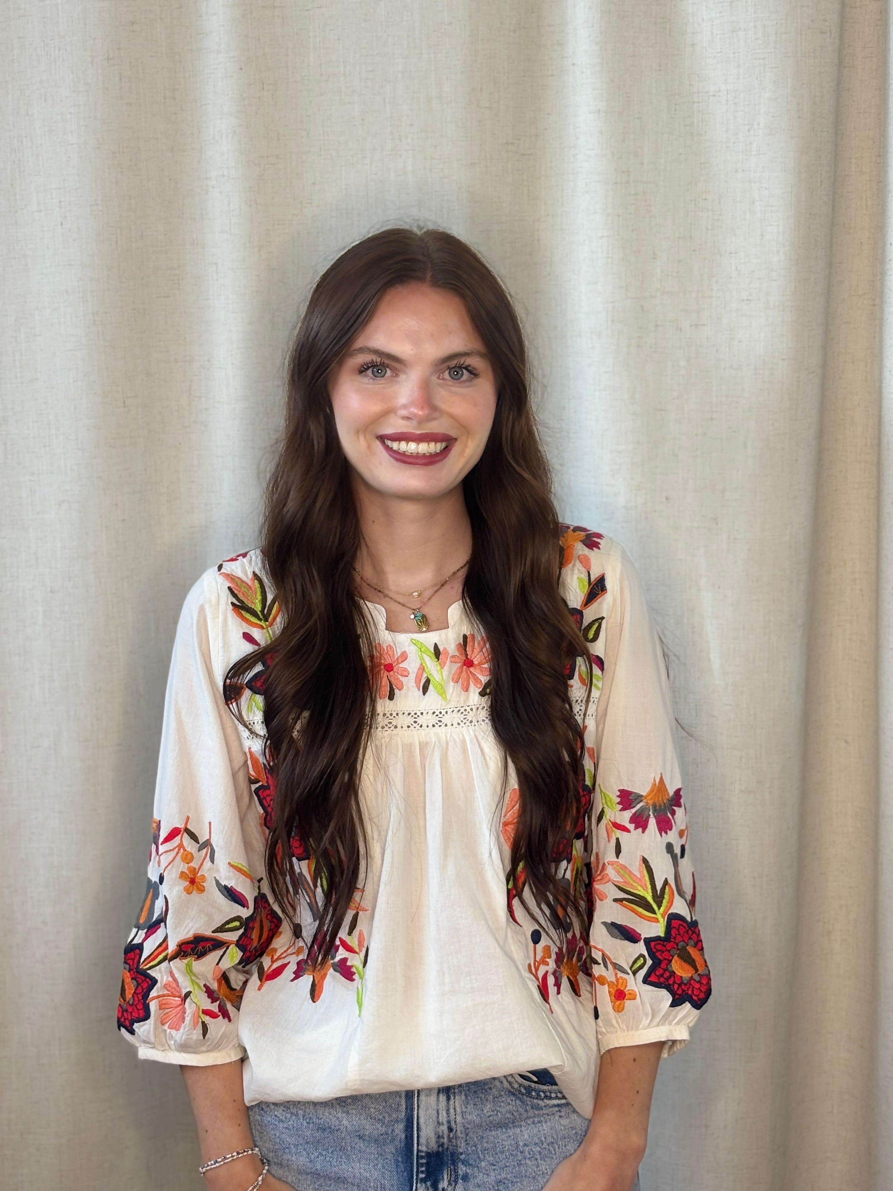 Woman wearing white cotton boho-luxe embroidered blouse with vibrant floral and leaf embroidery, lace trim detail, and 3/4 sleeves — front view, styled with denim jeans