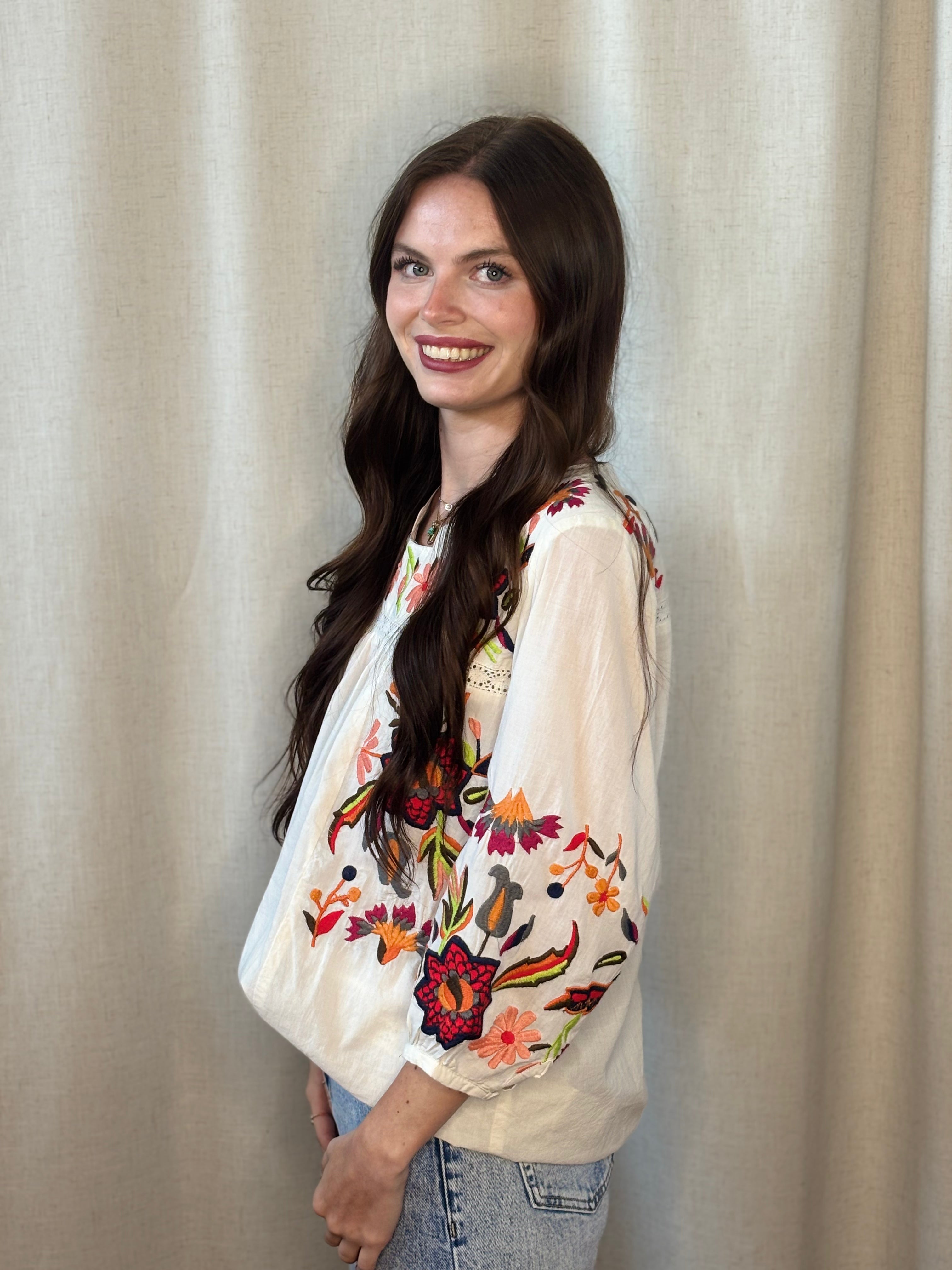 Side view of white artisan embroidered blouse with colorful floral embroidery on sleeves and chest, boho-luxe style, paired with blue jeans