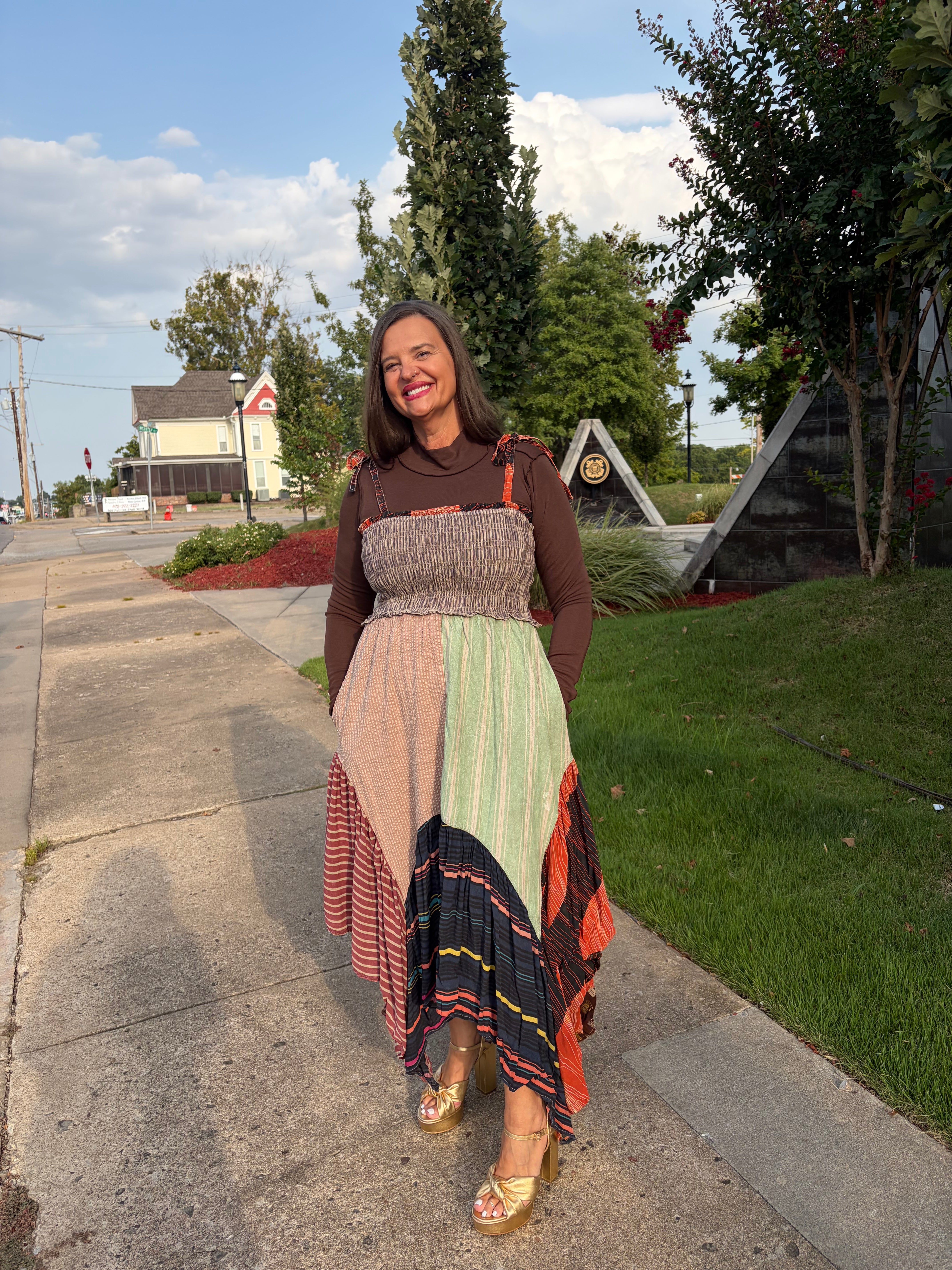 A woman stands on a sidewalk outdoors wearing a long-sleeve patchwork maxi dress with a smocked bodice and a multicolor striped skirt, paired with gold flat shoes.