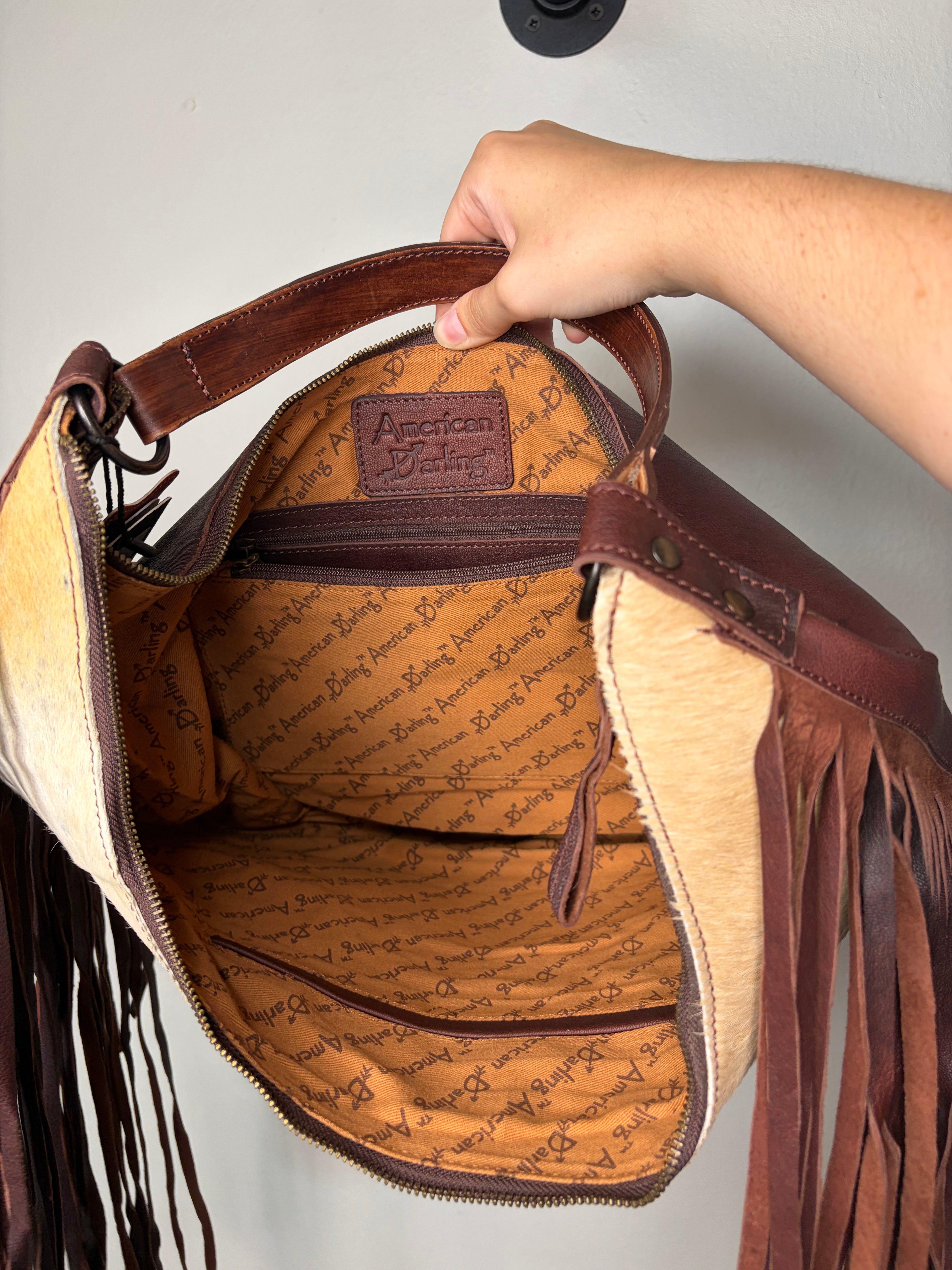 An open handbag with a tan and brown cowhide exterior, dark brown leather fringe, and a tan patterned interior lining. A hand is holding the top handle, and the brand name 'American Galley' is visible on the interior label.