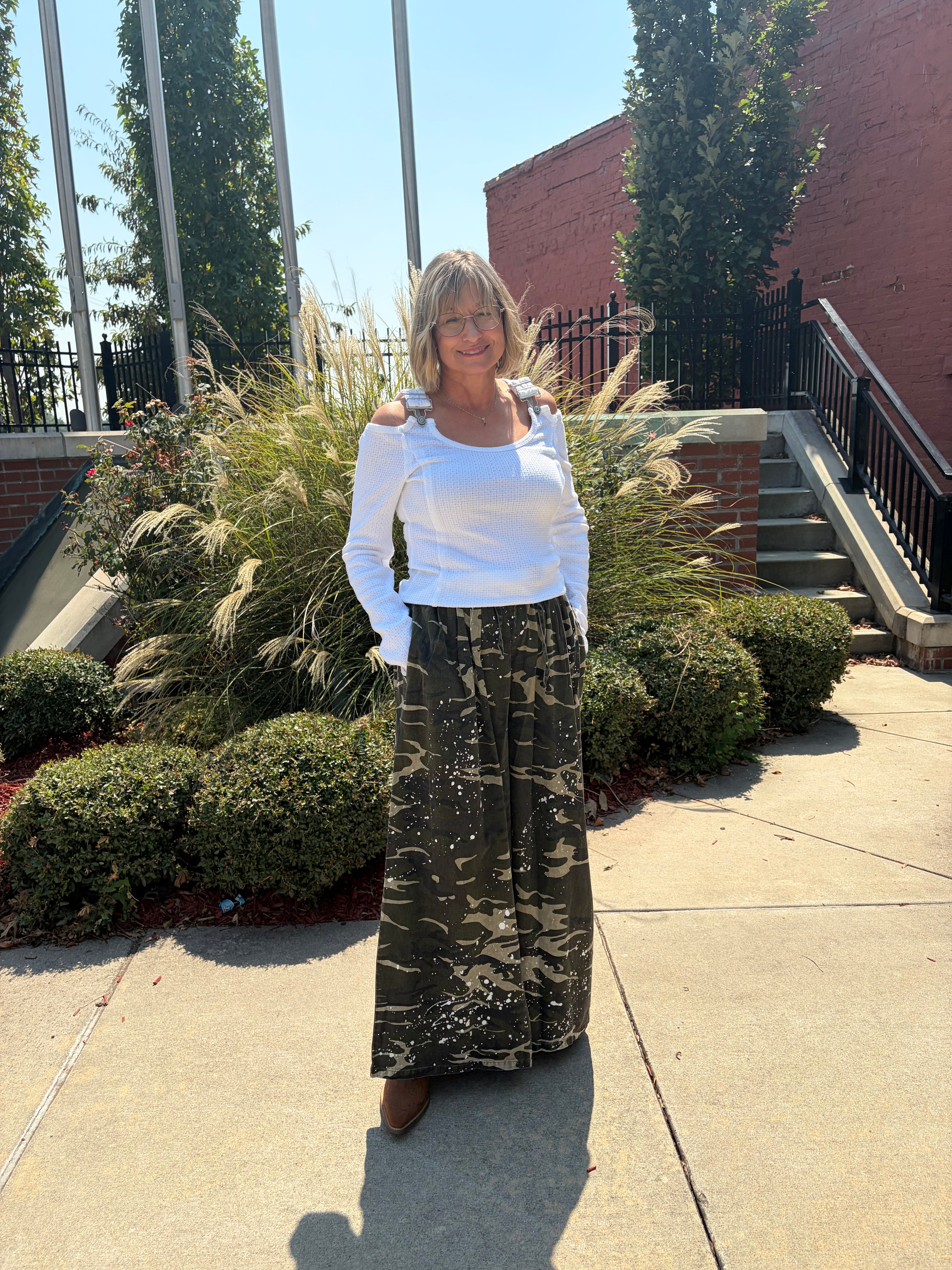 A woman wearing a white off-the-shoulder long-sleeve top and a dark green camouflage maxi skirt, standing outdoors on a concrete walkway with landscaping and a brick building in the background.