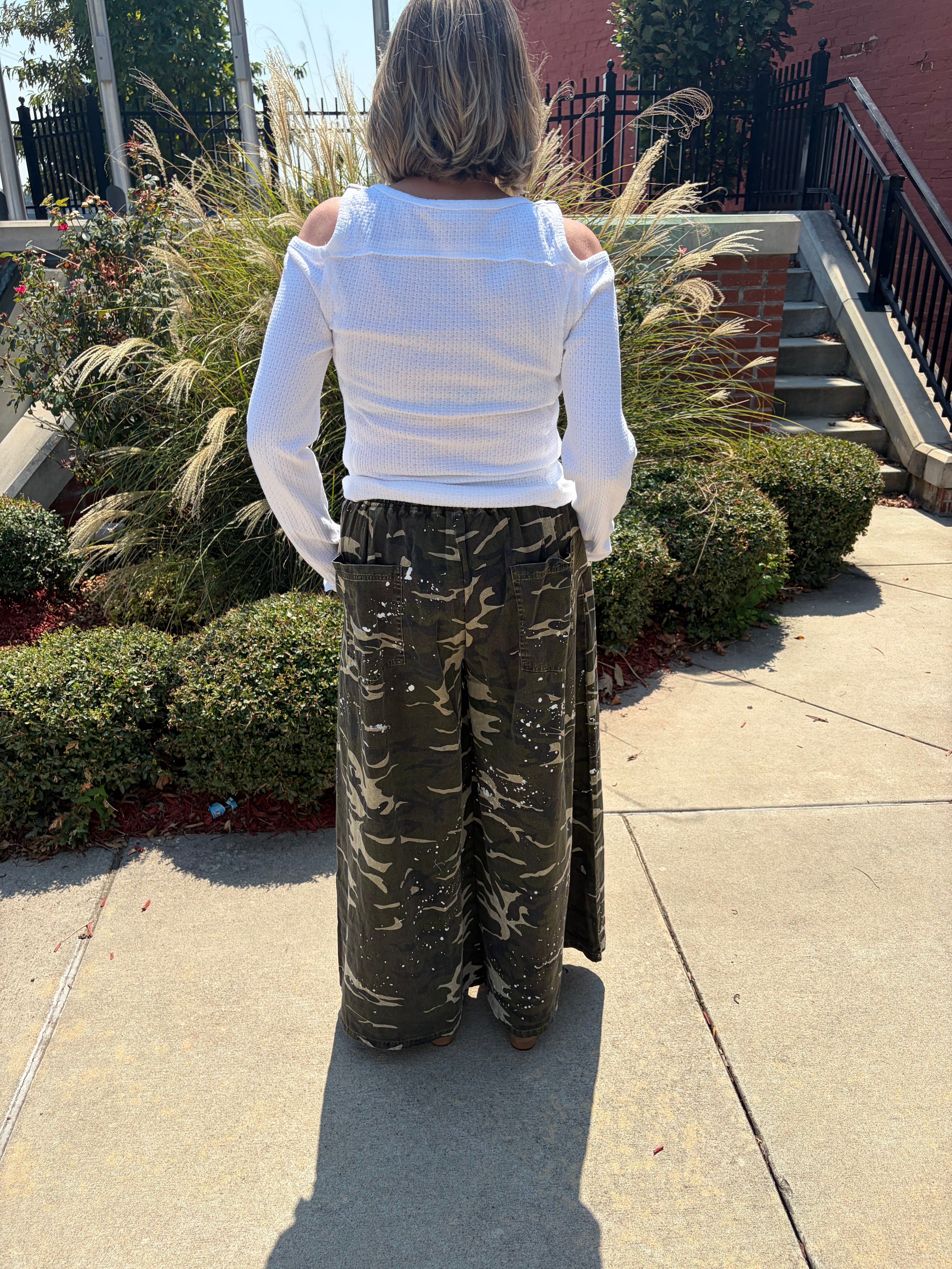 A woman stands outdoors on a sidewalk, viewed from behind, wearing a white long-sleeve top with shoulder cutouts and wide-leg camouflage pants.