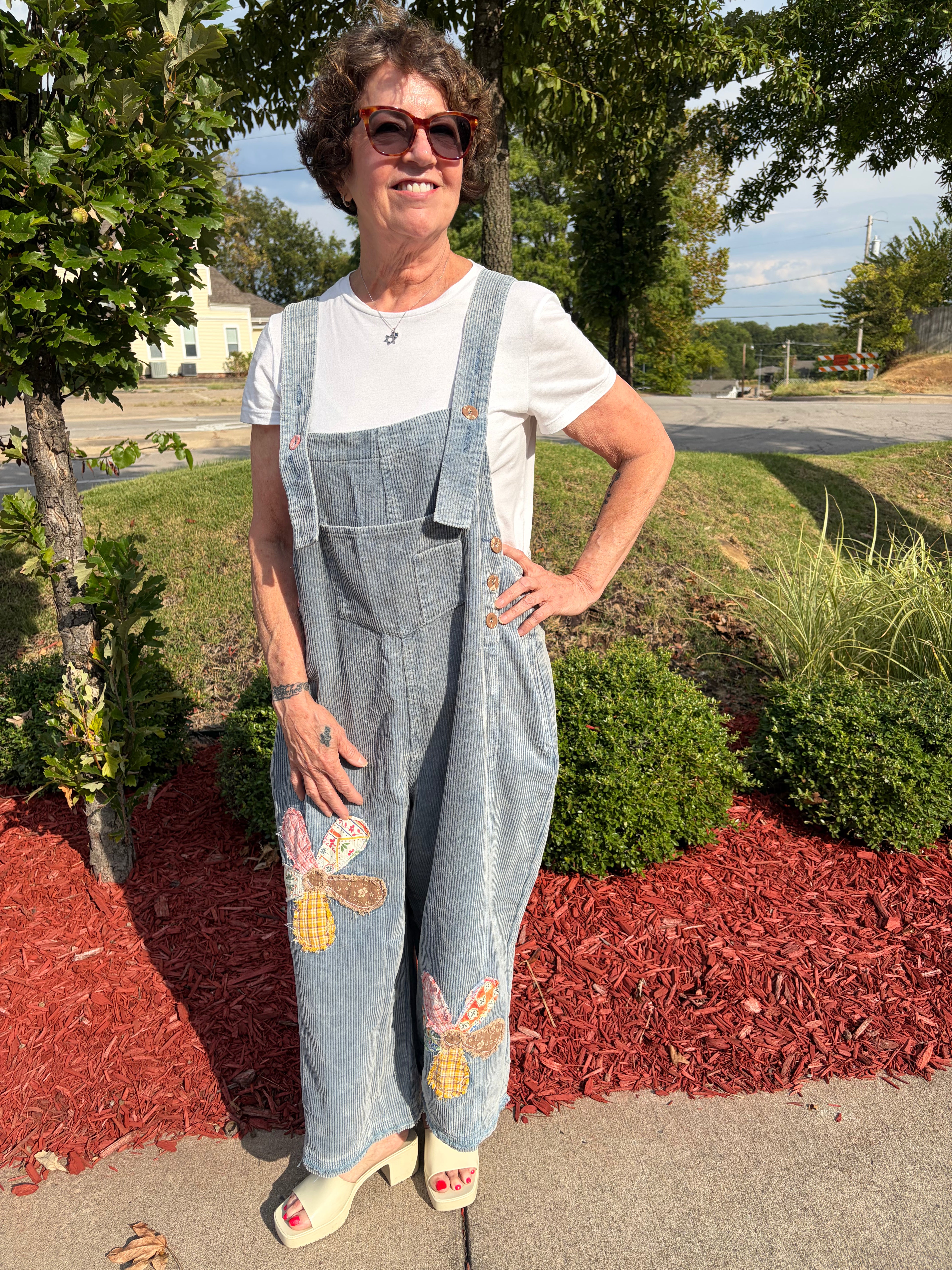 front full-length view of woman smiling wearing denim blue Flower Patch Corduroy Wide-Leg Overalls with white tee and white sandals, showing floral patchwork detail on both legs