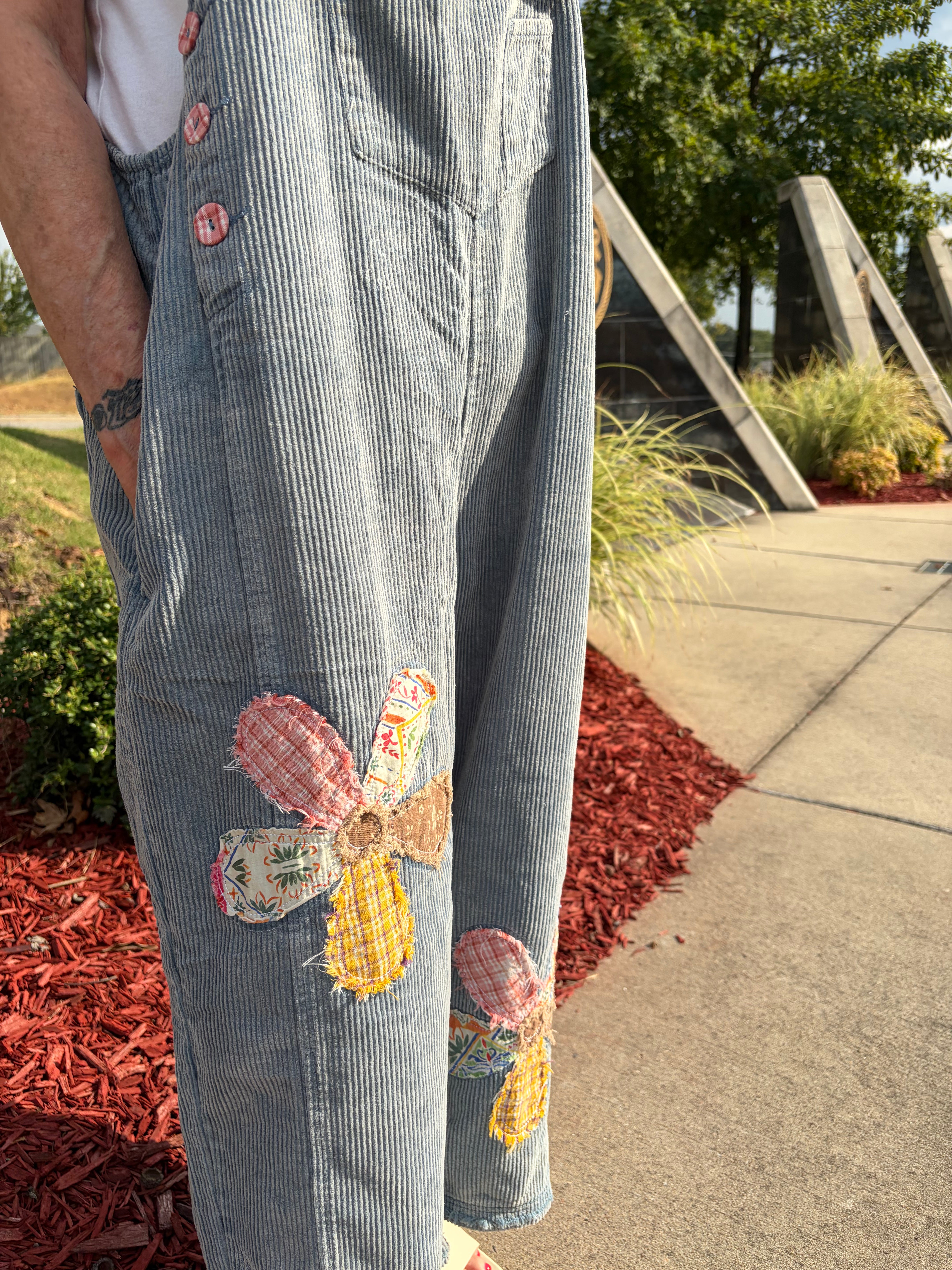 A close-up of a person wearing light blue corduroy overalls with colorful patchwork flower appliqués on the legs, standing outdoors on a paved walkway with mulch and greenery in the background.