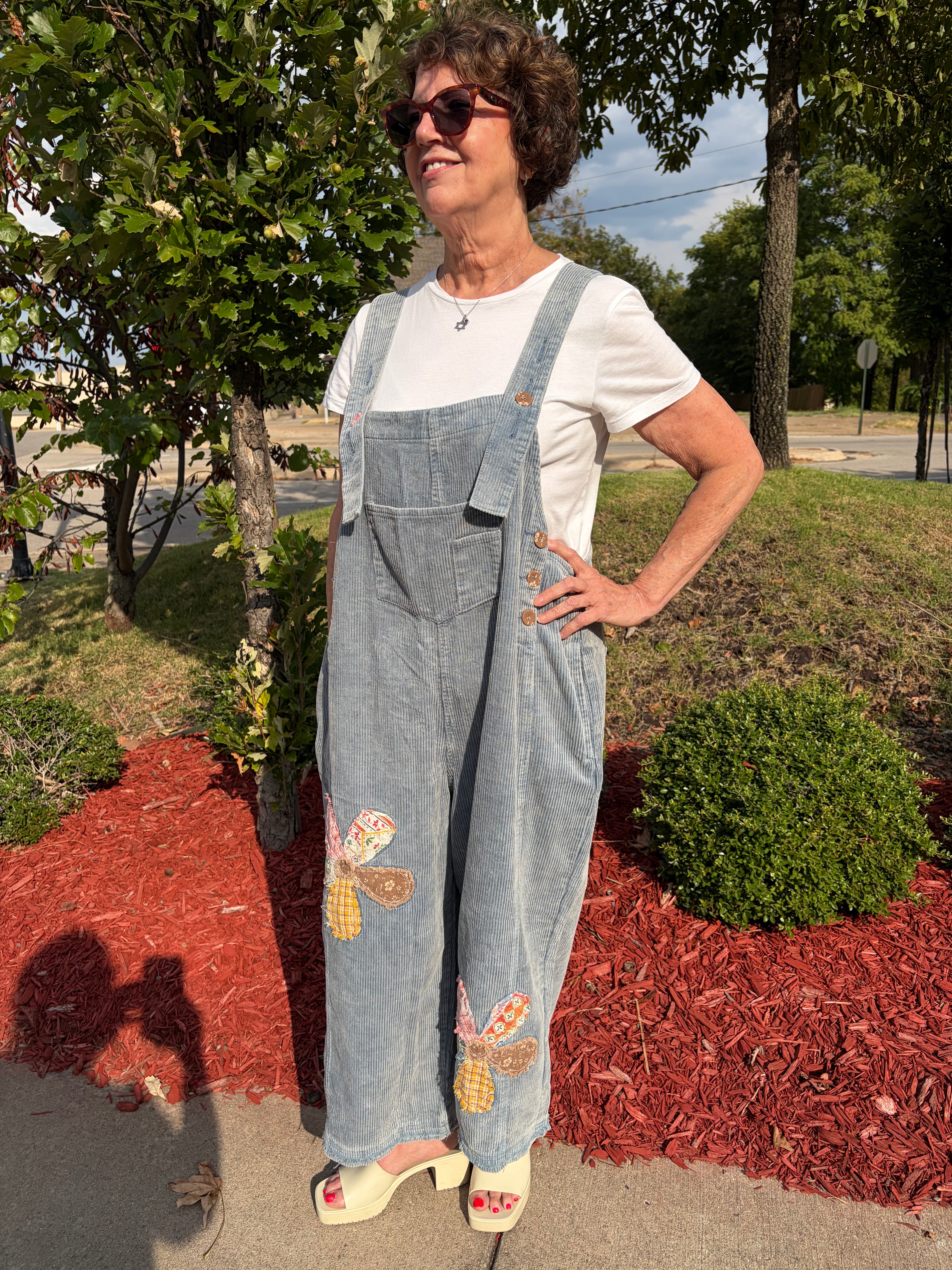 Front full-length view of woman smiling wearing olive green Flower Patch Corduroy Wide-Leg Overalls with white tee and white platform sandals, outdoor setting