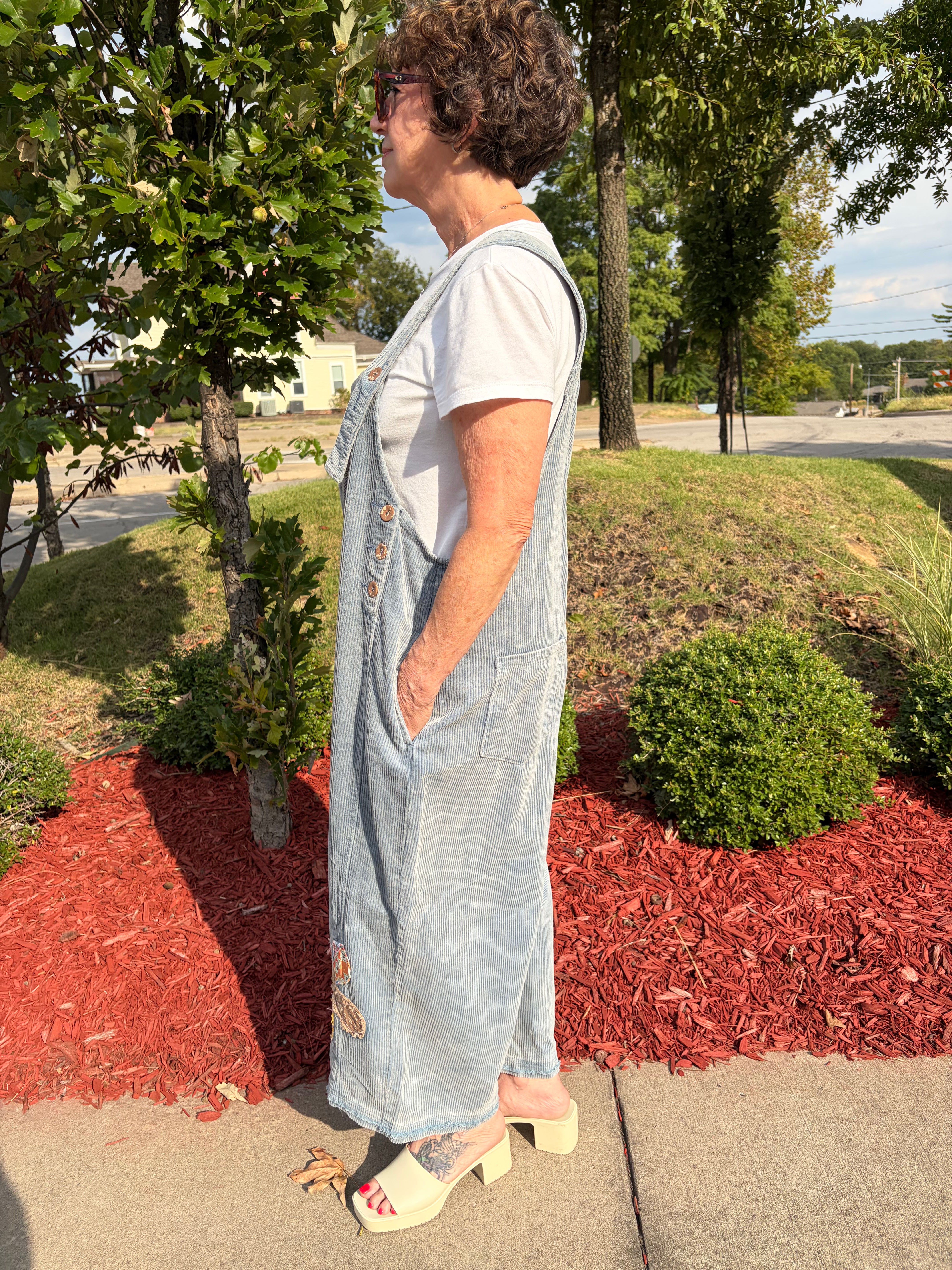 Close-up side view of olive green corduroy wide-leg overalls showing mixed-print floral patchwork appliqué on leg, side button closure, and front pocket detail