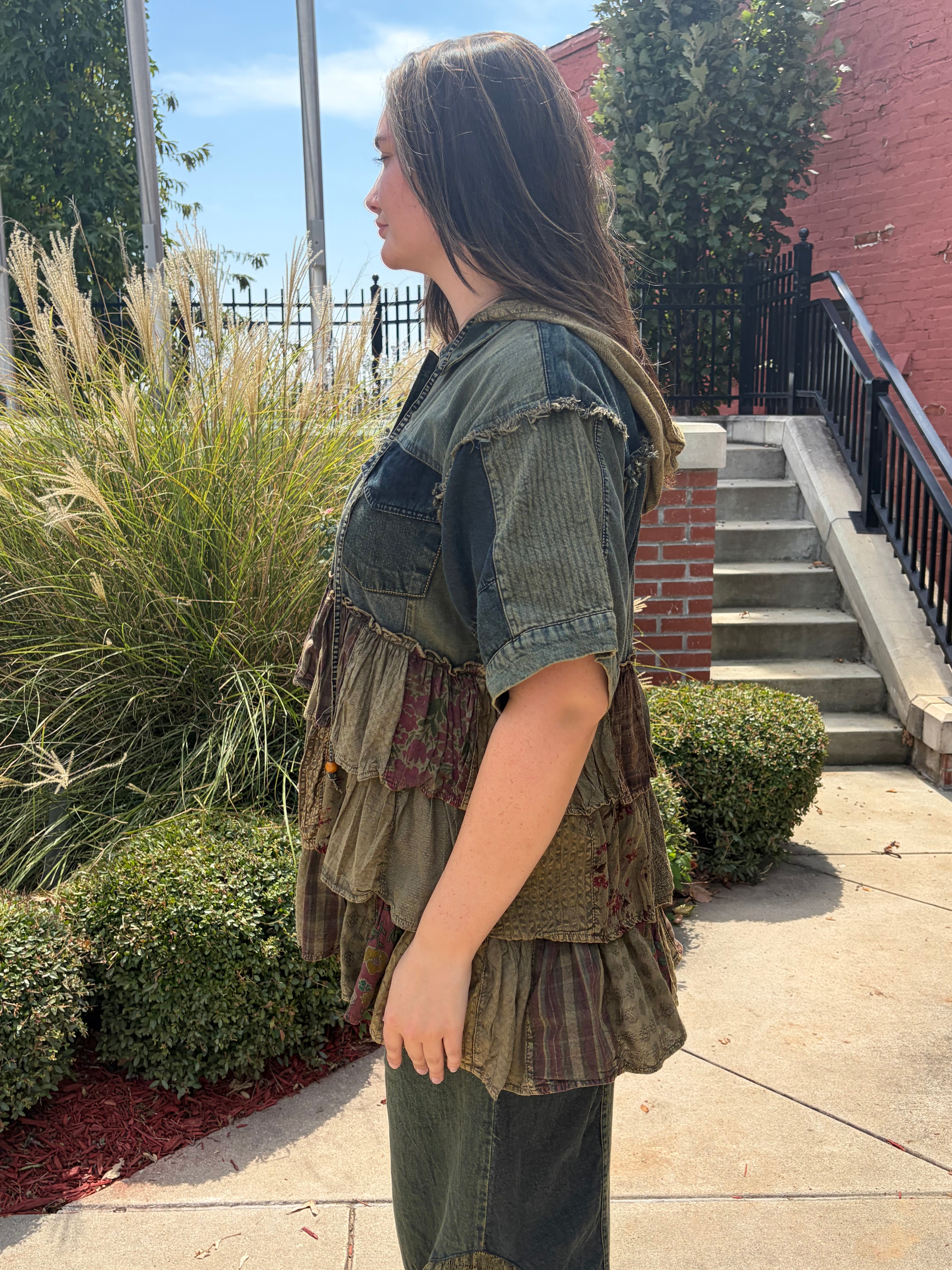 A woman wearing a handmade, multi-layered patchwork vest with denim and earthy fabric details, standing outdoors on a concrete walkway next to green plants and a brick staircase.
