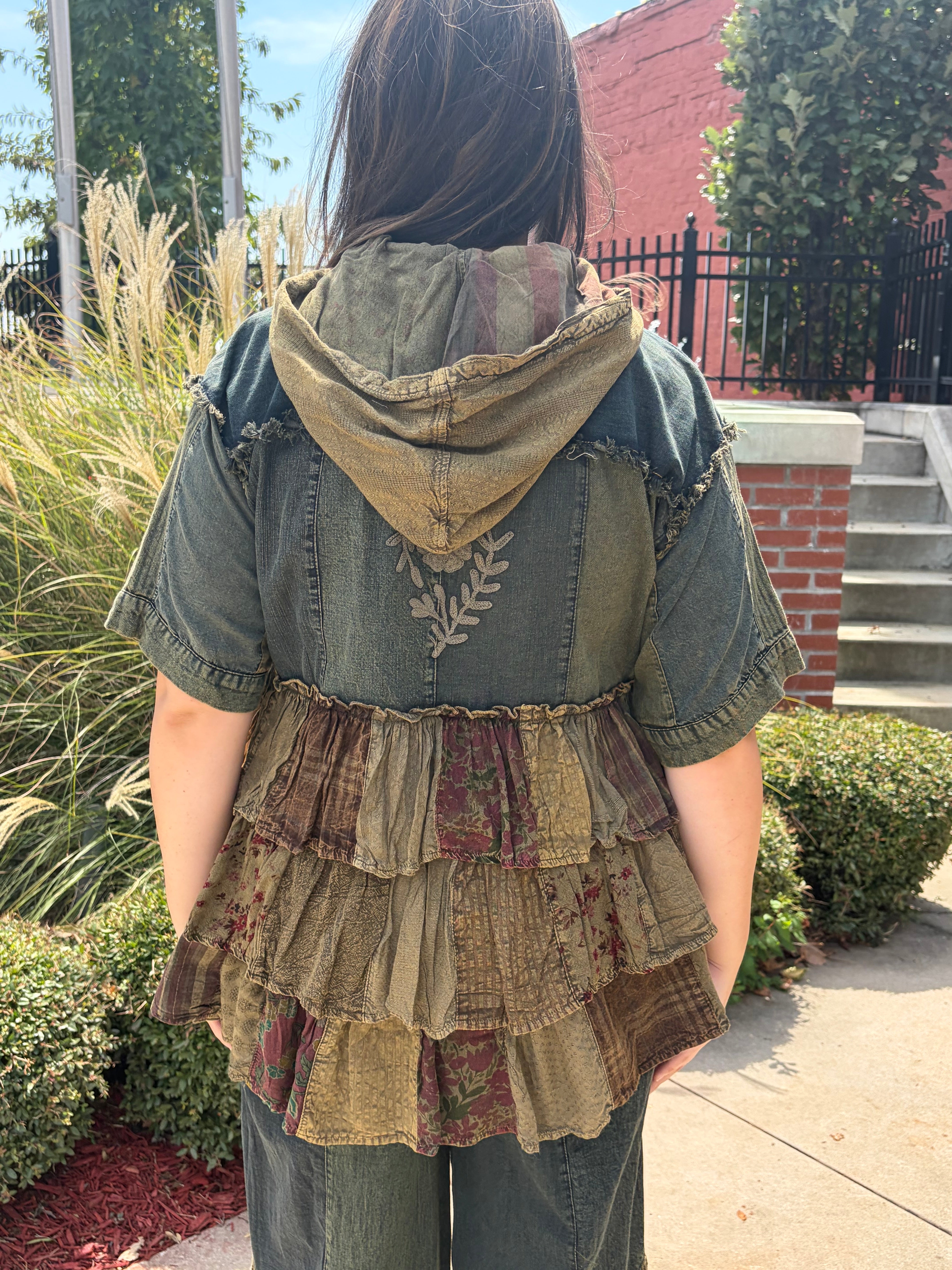 A person viewed from behind wearing a patchwork-style hooded top with a denim-look back panel and layered ruffle skirt in earthy olive, brown, and burgundy tones, photographed outdoors on a sidewalk with shrubs and a brick wall in the background.