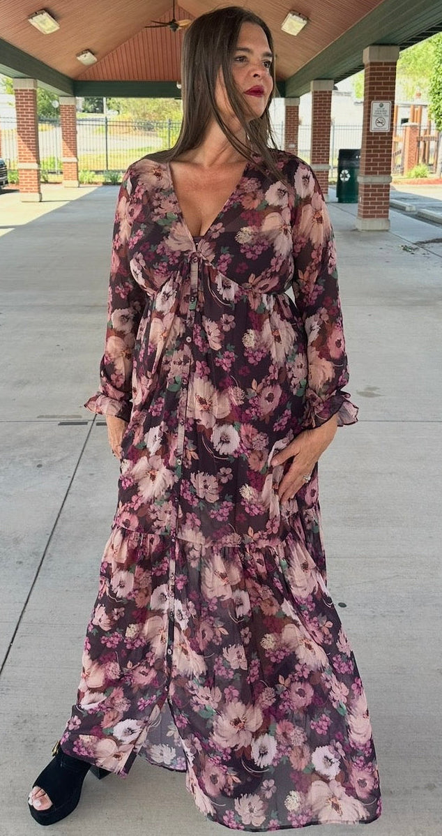 A woman wearing a long-sleeve, floor-length floral maxi dress with a V-neckline, standing under a covered walkway.