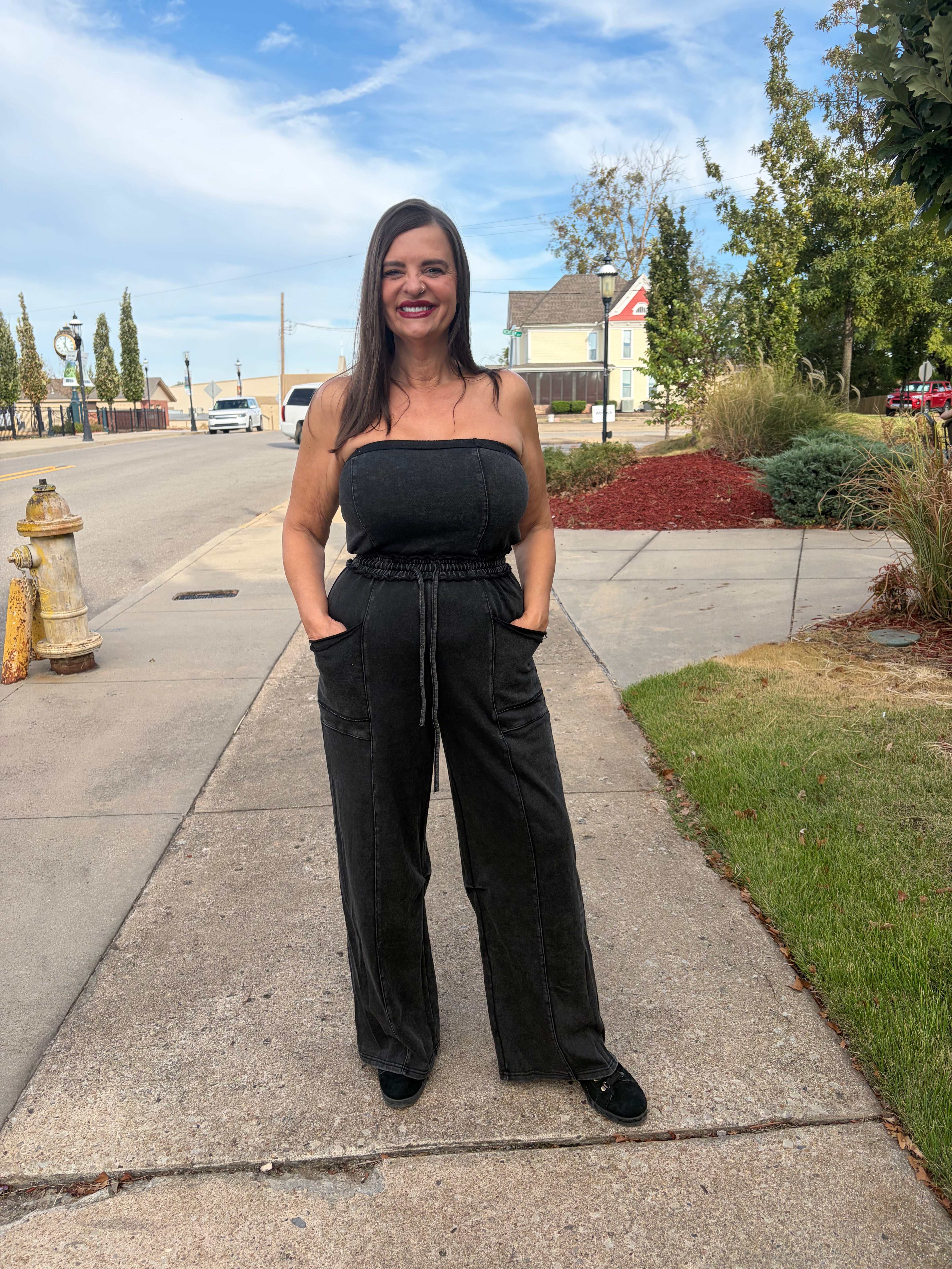 A woman stands on a sidewalk outdoors wearing a black strapless jumpsuit with wide legs and black shoes. The background shows a street, hydrant, grass, trees, and a house under a blue sky.
