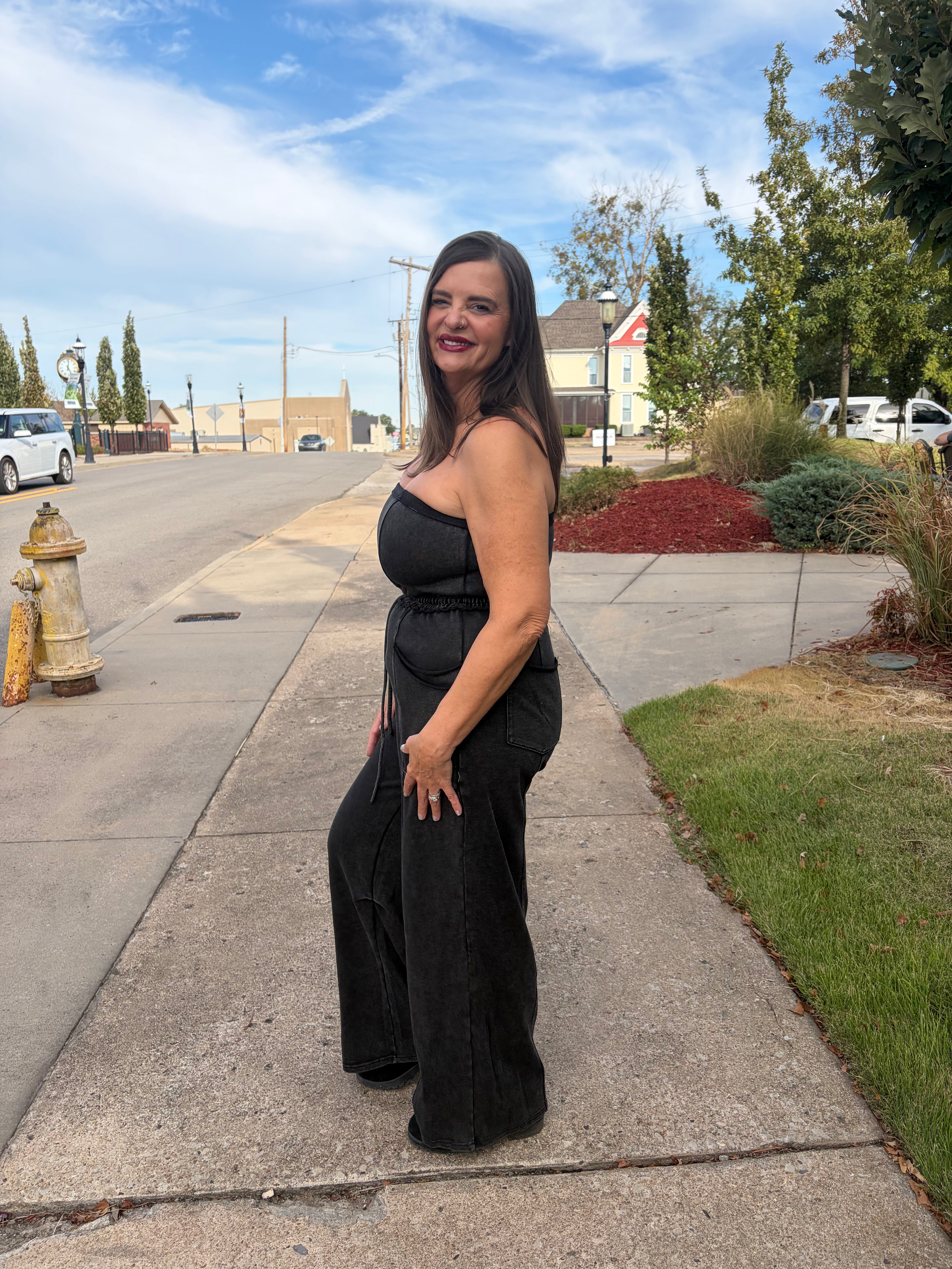 A woman wearing a black strapless jumpsuit with wide-leg pants stands on a suburban sidewalk, looking back over her shoulder. The background shows a street, trees, and a blue sky.