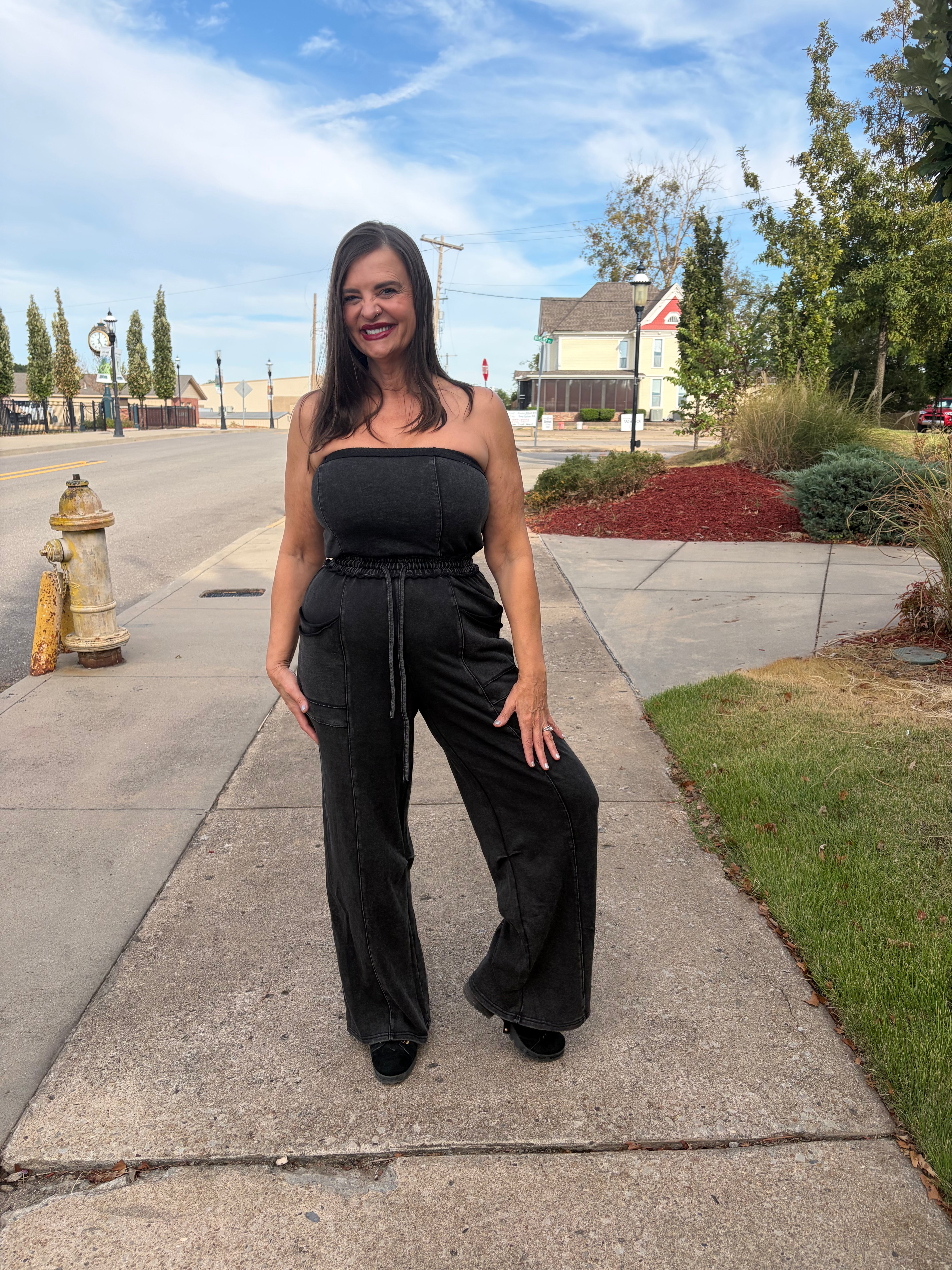 A woman wearing a black strapless jumpsuit with wide-leg pants stands on a concrete sidewalk outdoors. The background shows a street, hydrant, grass, trees, and a house under a blue sky with clouds.