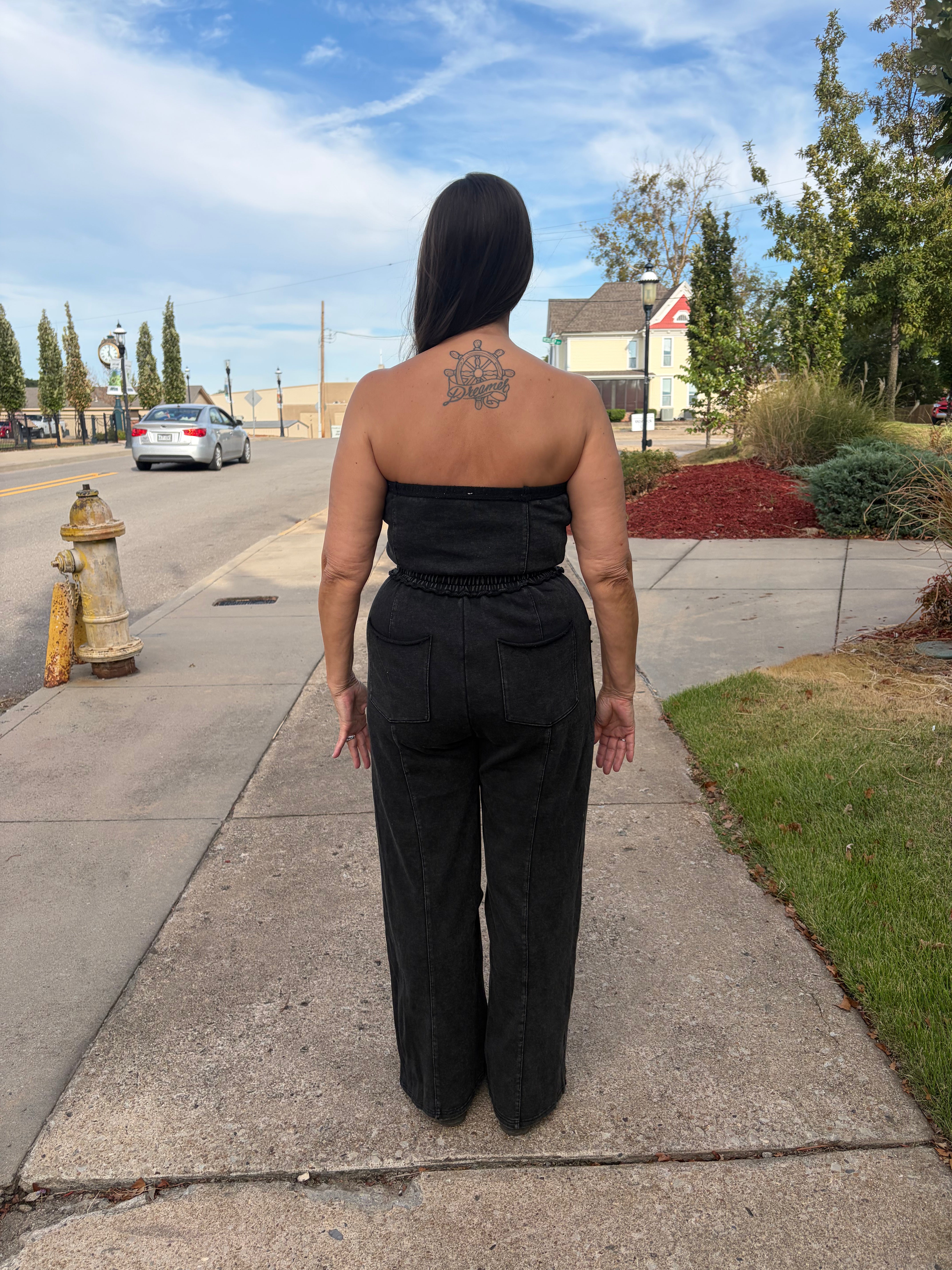 A woman facing backwards wearing a black strapless jumpsuit with wide-leg pants stands on a concrete sidewalk outdoors. The background shows a street, hydrant, grass, trees, and a house under a blue sky with clouds.