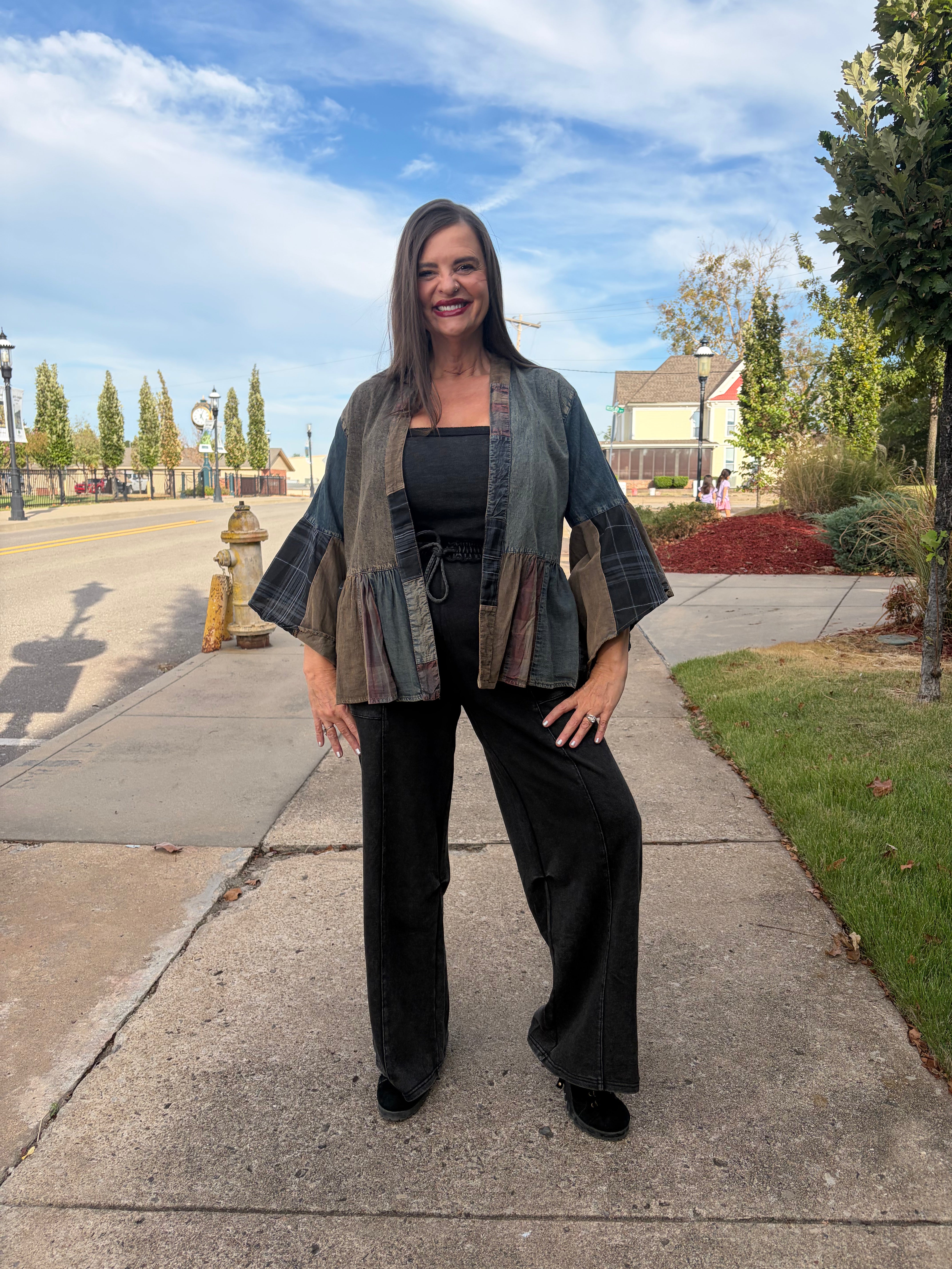A woman stands on a suburban sidewalk wearing a patchwork-style open-front jacket with wide sleeves, a black top, and black wide-leg pants.
