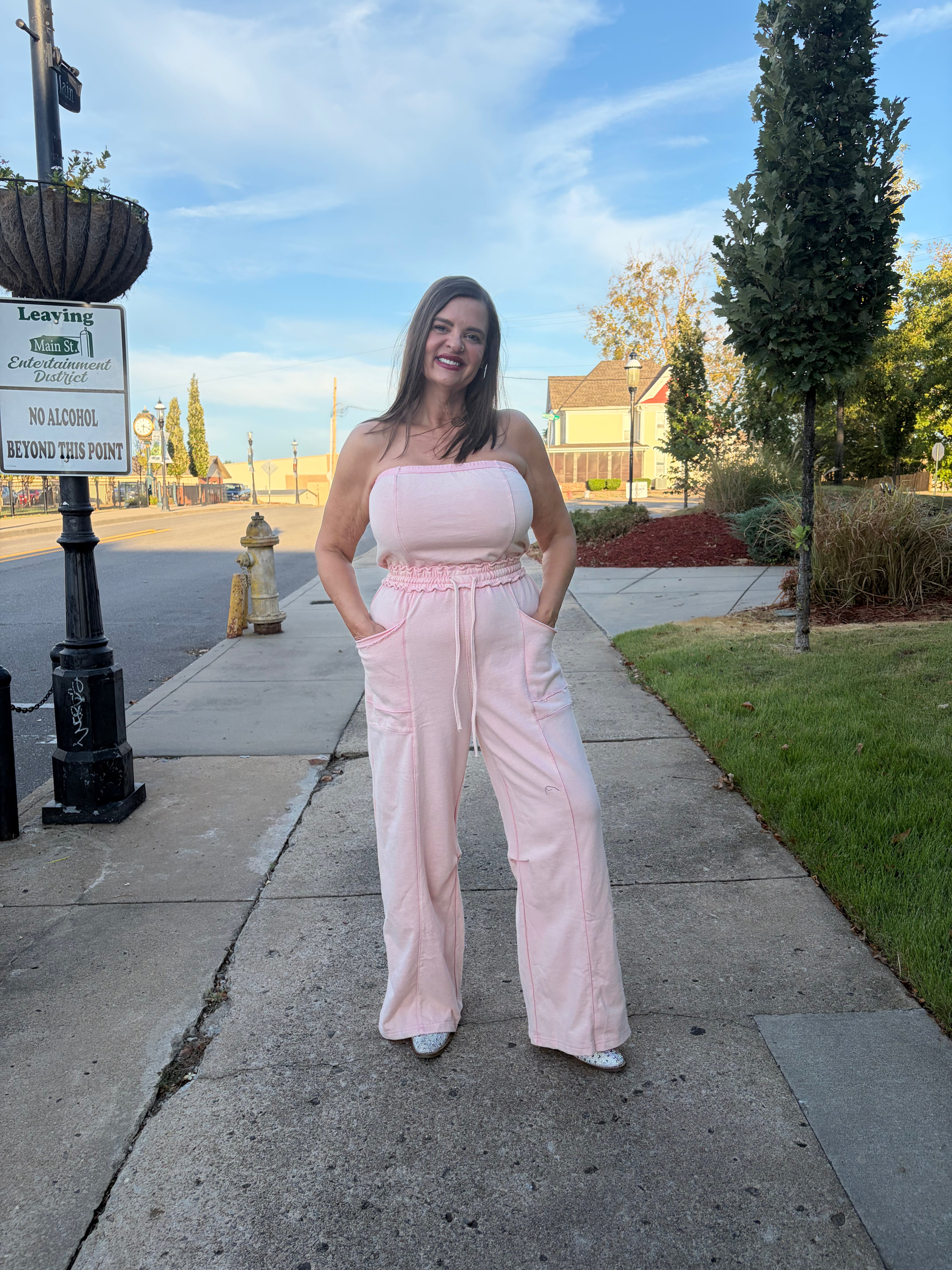 A woman stands on a sidewalk outdoors wearing a light pink strapless jumpsuit with wide legs and a drawstring waist. The background shows a street, grass, trees, and a blue sky.