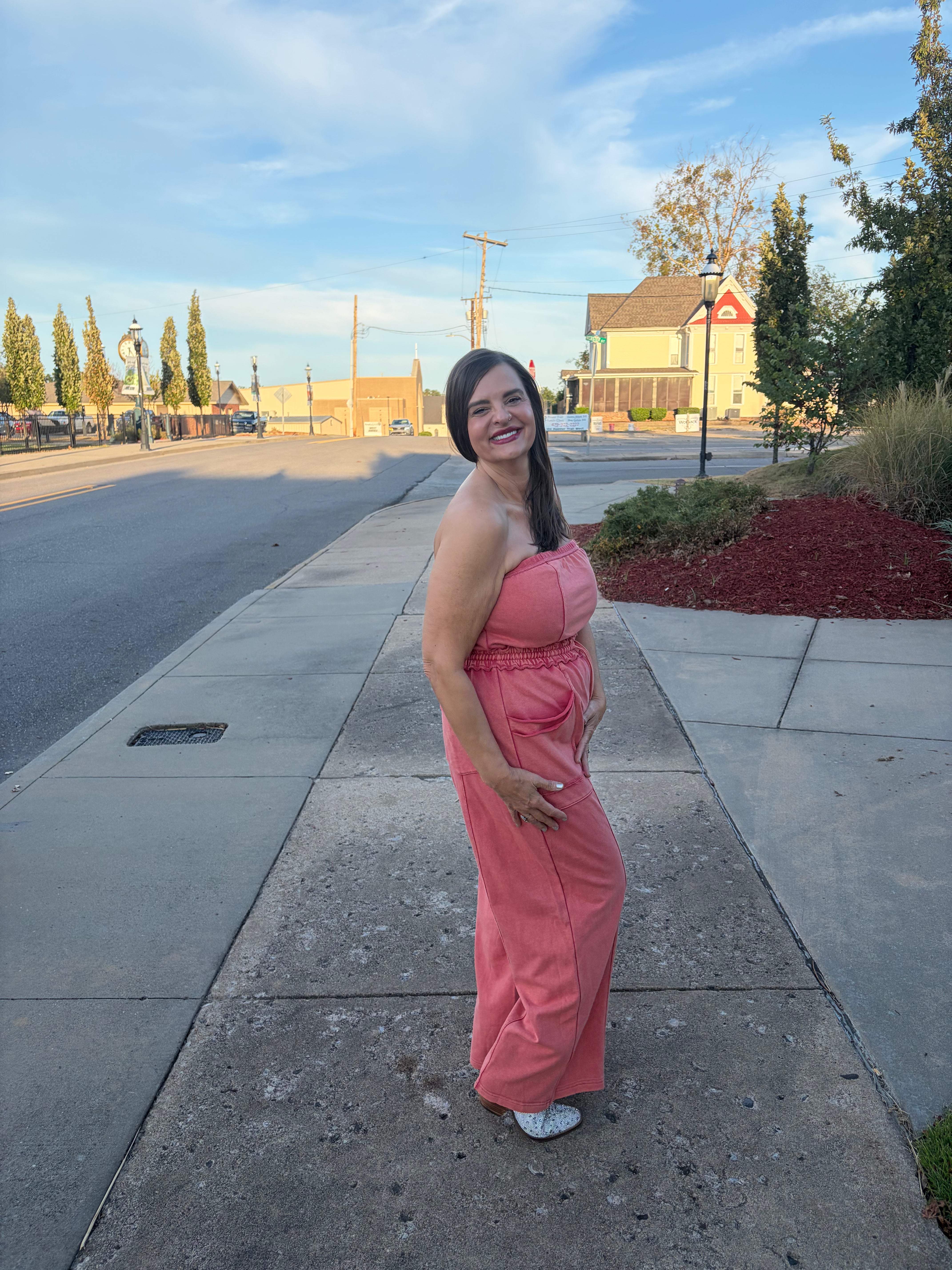 A woman wearing a strapless pink maxi dress stands on a concrete sidewalk outdoors, looking back over her shoulder. The background shows a street, trees, and a building under a blue sky.