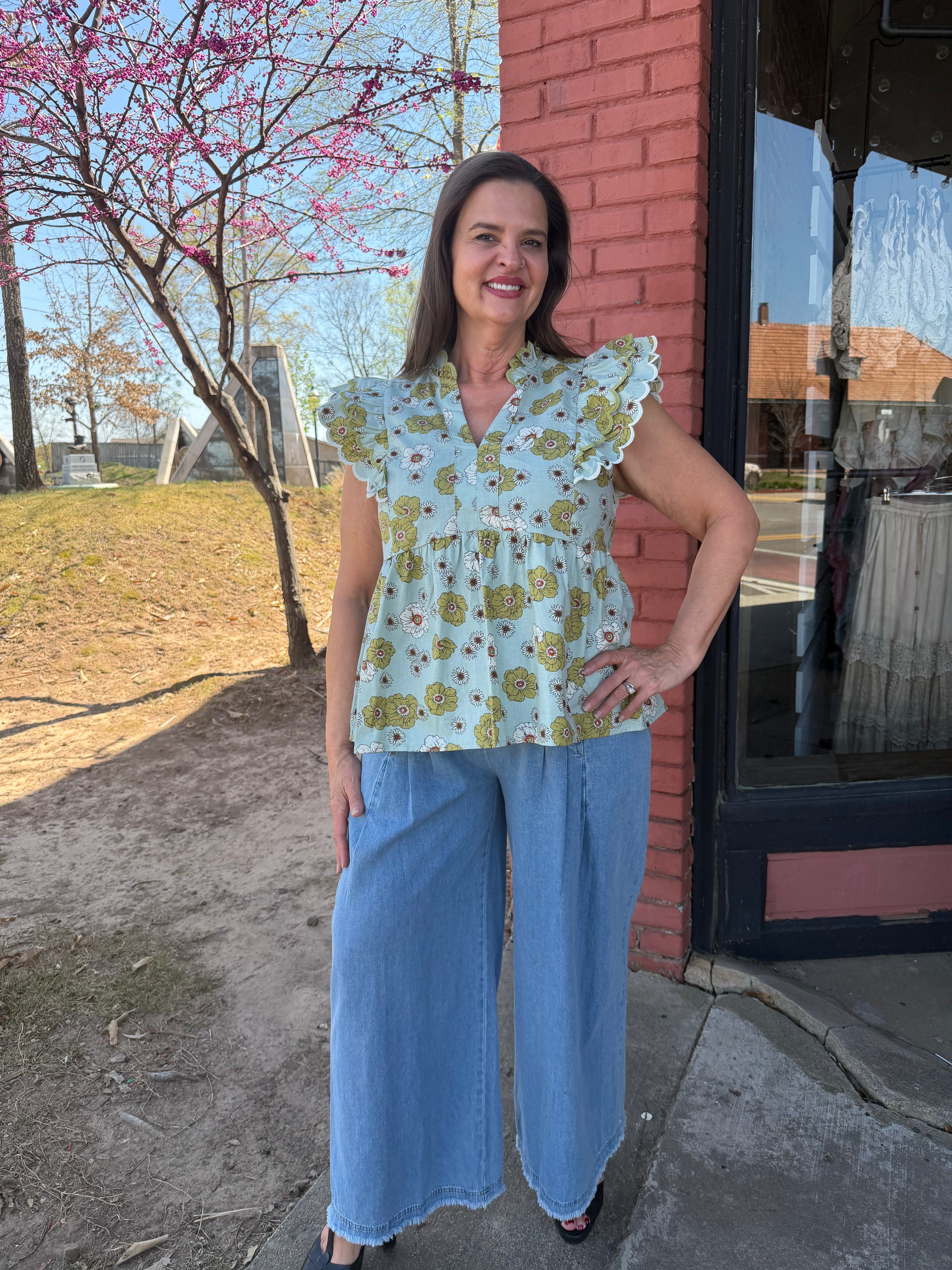 Woman wearing a floral top and blue pants standing outside a building with pink brick walls.