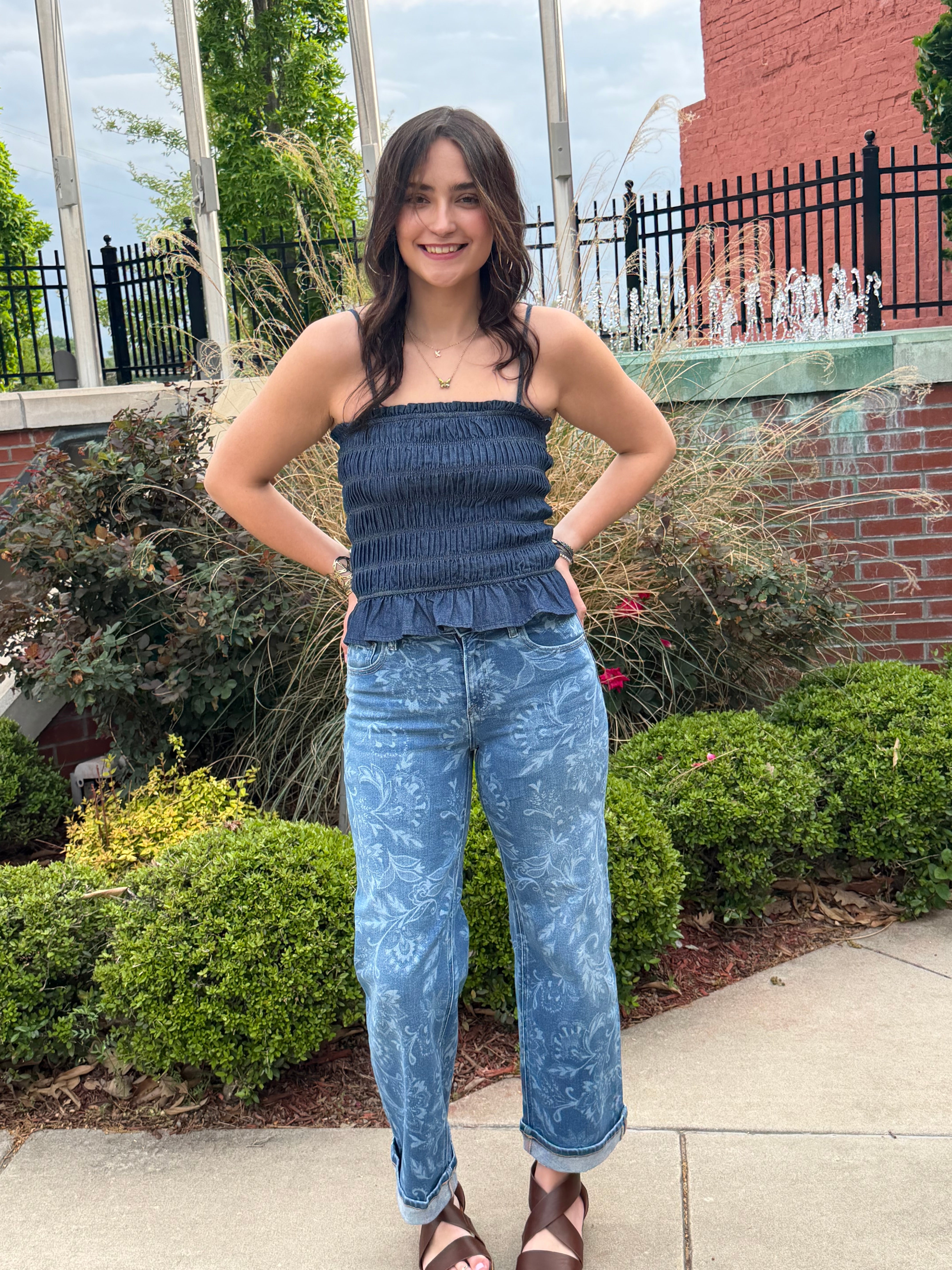 Person wearing a blue top and jeans standing outdoors with greenery and a building in the background